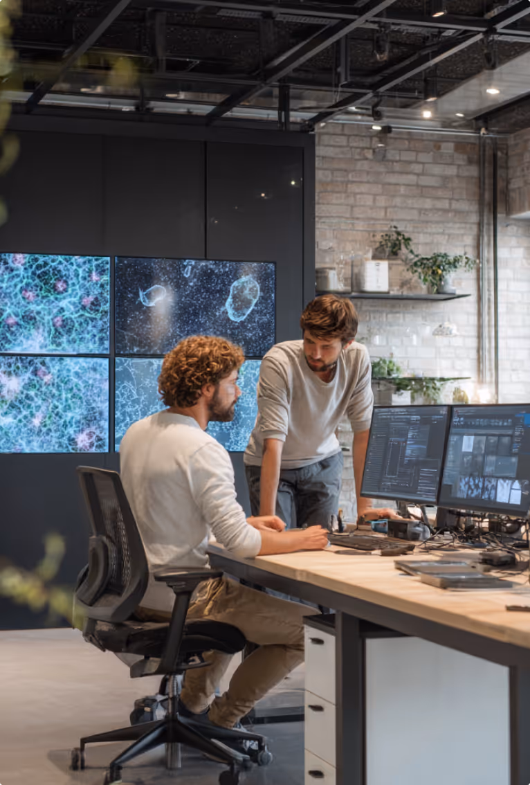 Two men collaborating at a desk with multiple computer monitors displaying data and scientific images in a modern office.