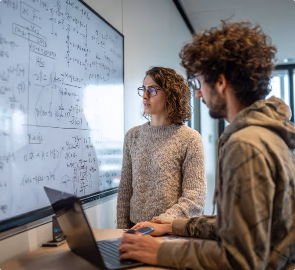 Two people standing by a whiteboard covered with mathematical equations, one using a laptop and the other looking at the board.