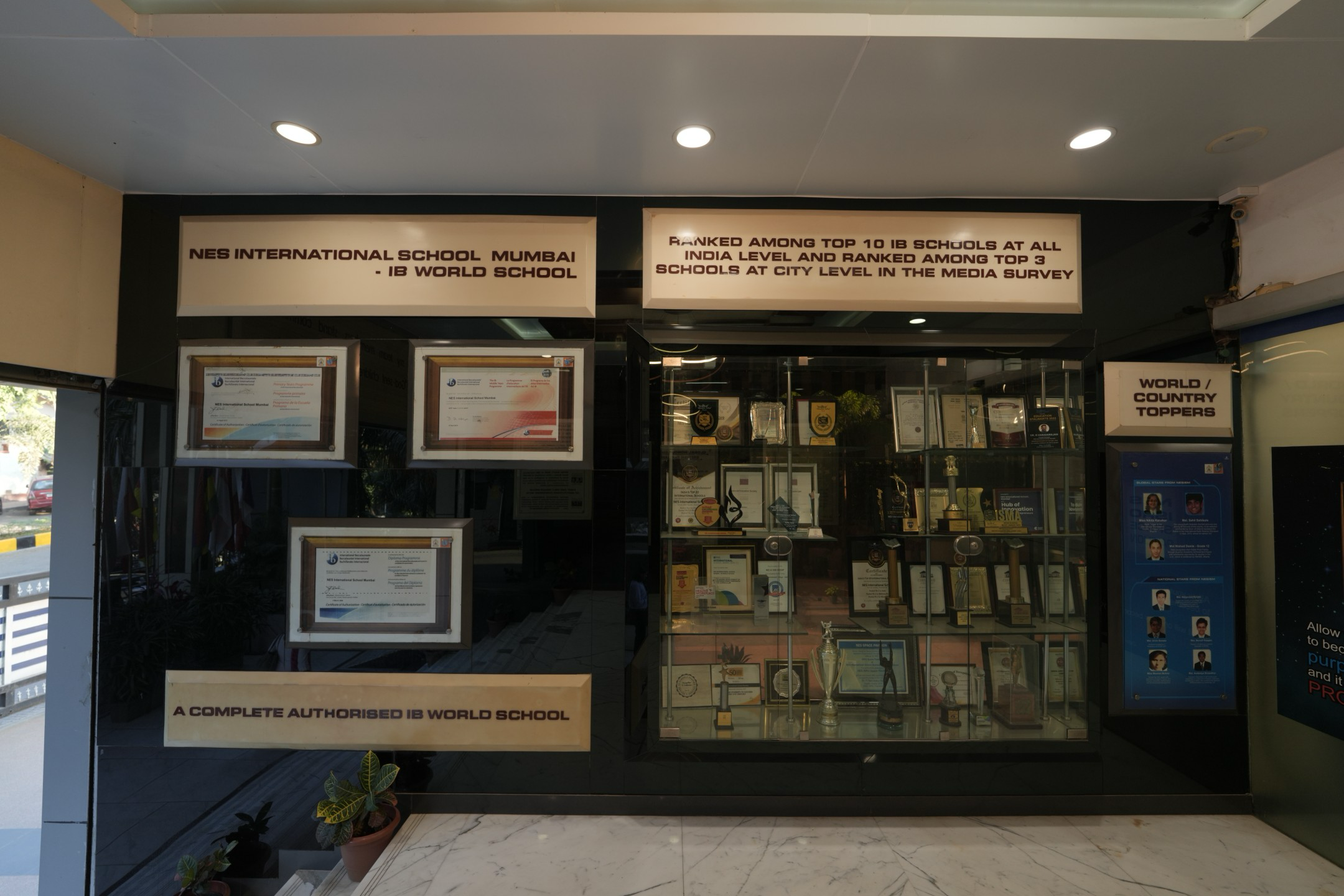 Display wall at NES International School Mumbai showing framed certificates, awards, trophies, and photos of world and country toppers.