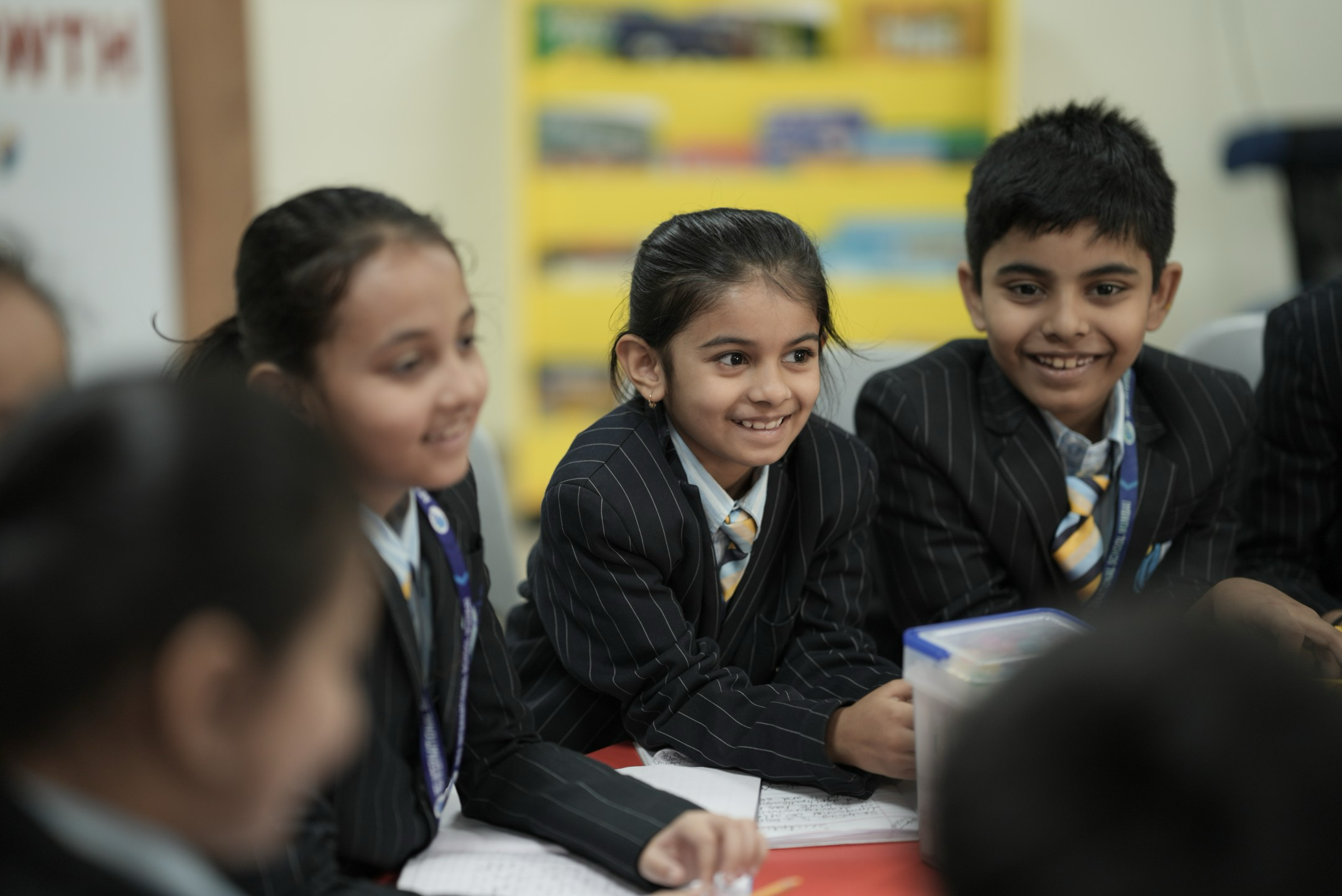 Group of smiling schoolchildren in striped uniforms sitting at a table with notebooks and a container.