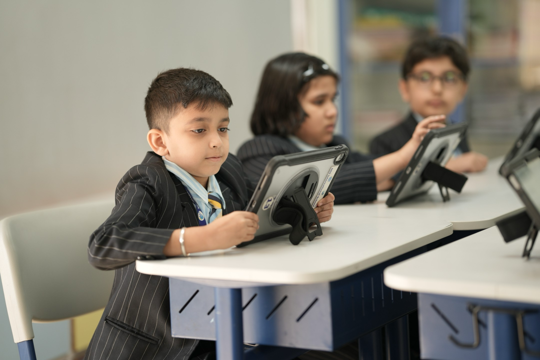 Young students in school uniforms seated at desks, using tablet devices with protective cases and stands.