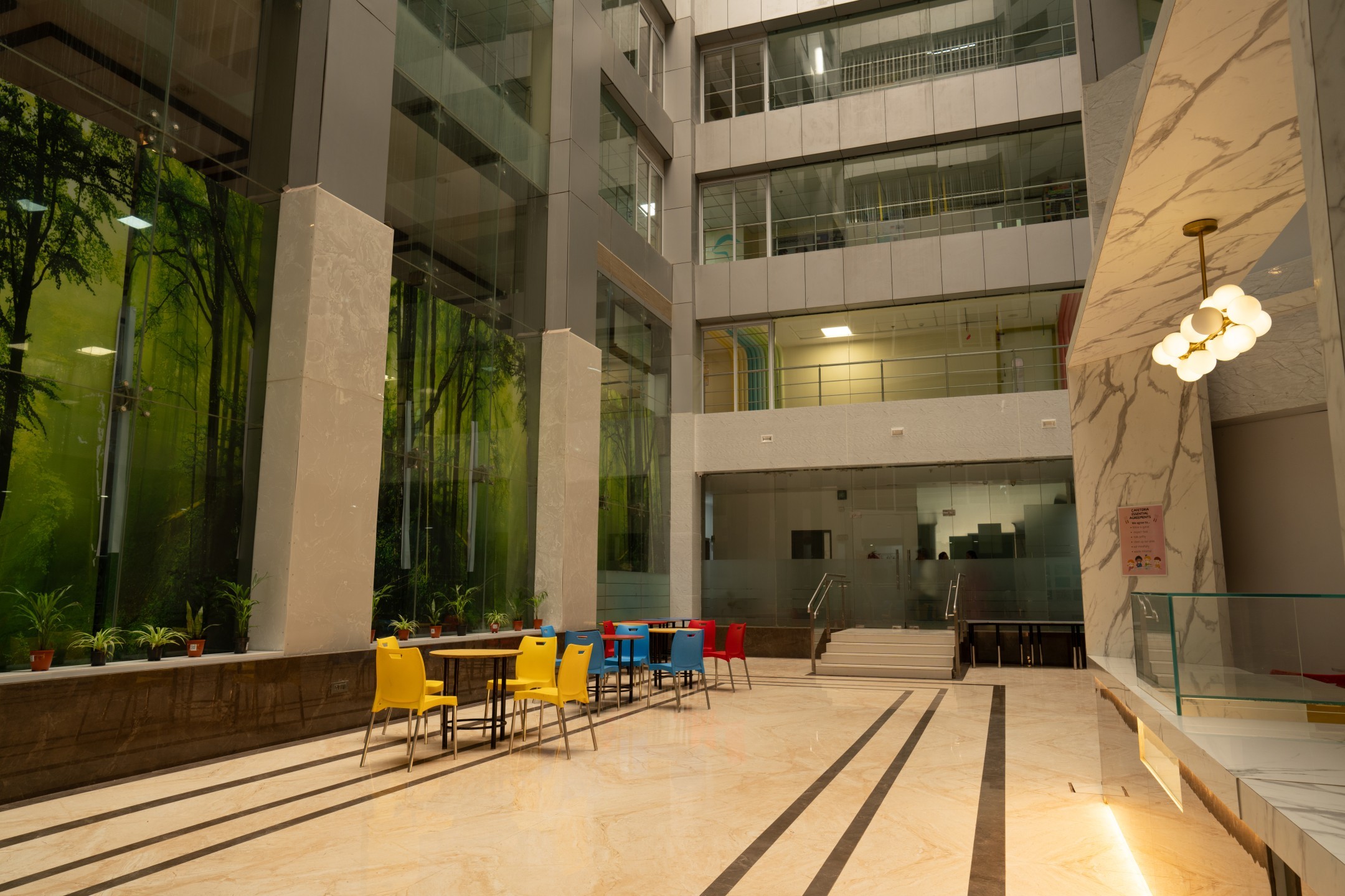 Spacious modern indoor seating area with yellow, blue, and red chairs around tables, large glass windows, and plants.