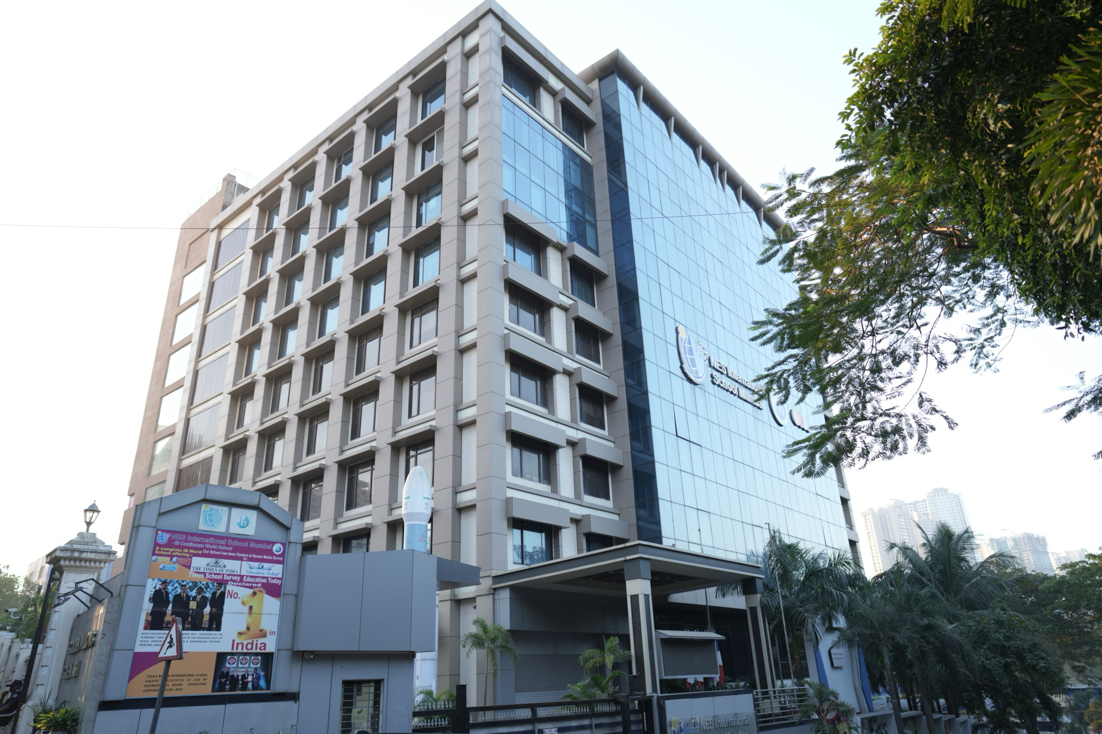 Modern multi-story building with glass facade labeled NES International School Mumbai, surrounded by trees and urban skyline.