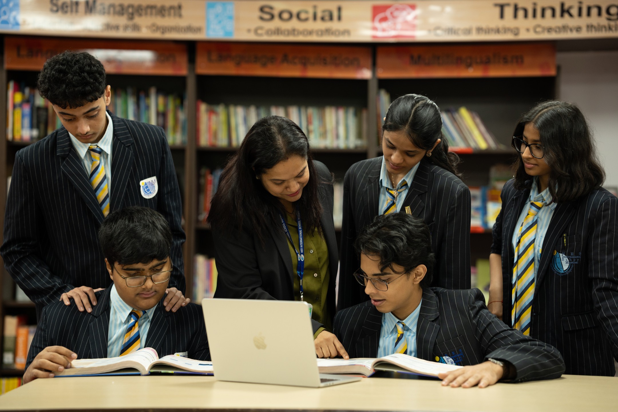 A teacher assisting five students in school uniforms with books and a laptop in a library setting.