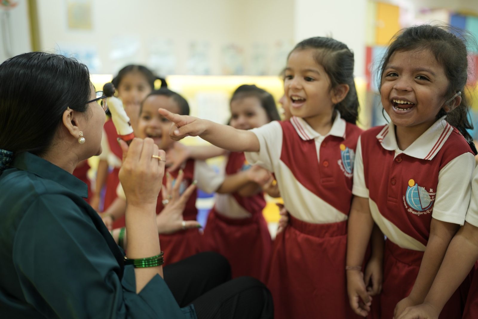 A teacher holding a puppet interacts with joyful young students wearing red and white uniforms in a classroom.