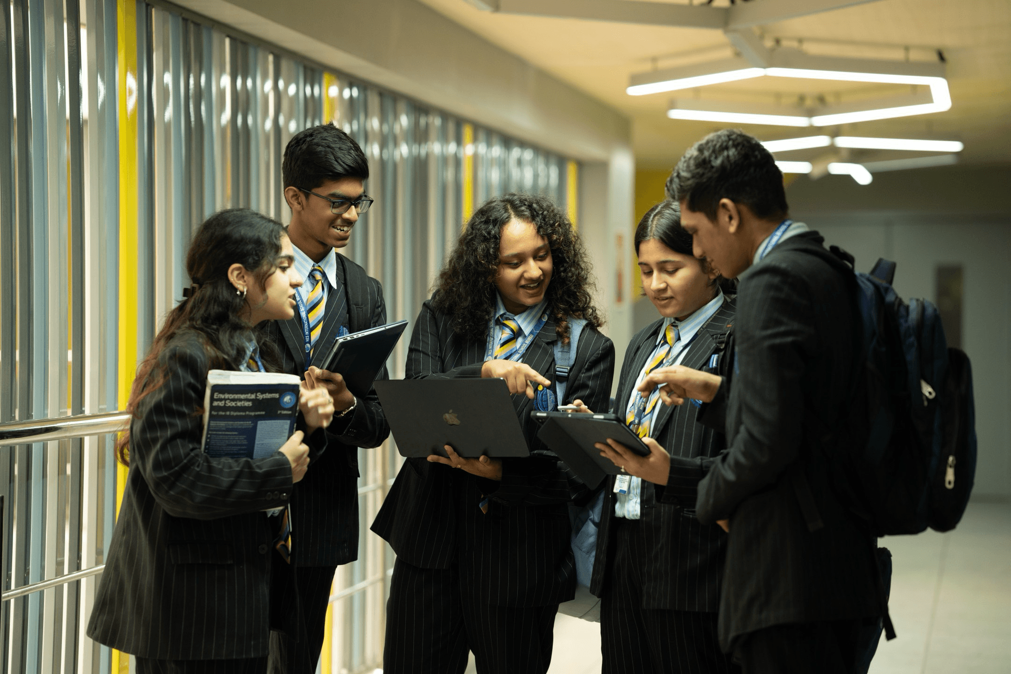 A female student wearing a striped blazer and yellow-blue tie attentively sitting at a desk with a laptop in a classroom.