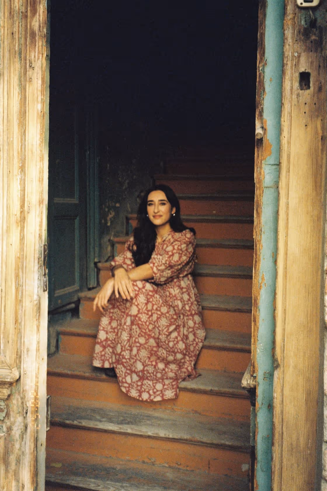 Woman with long dark hair wearing a patterned dress, sitting on rustic wooden stairs framed by weathered door trim.