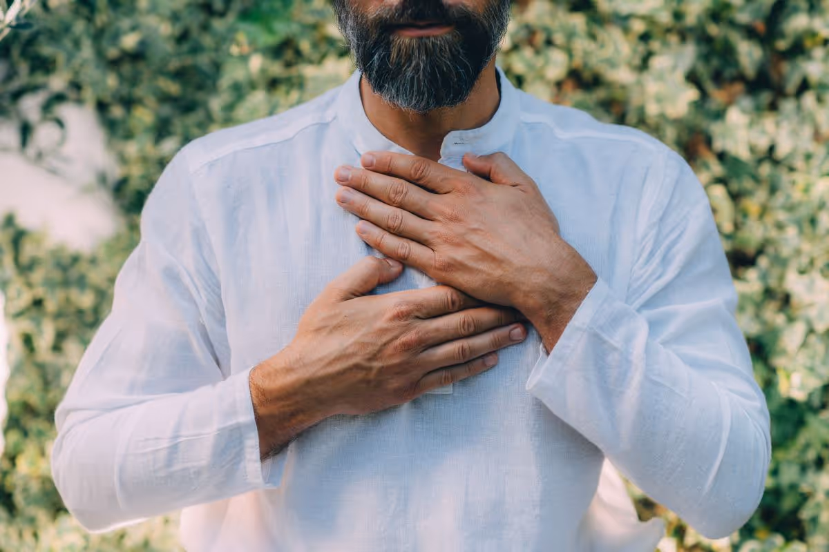Man with a beard wearing a white shirt placing hands over his heart outdoors.