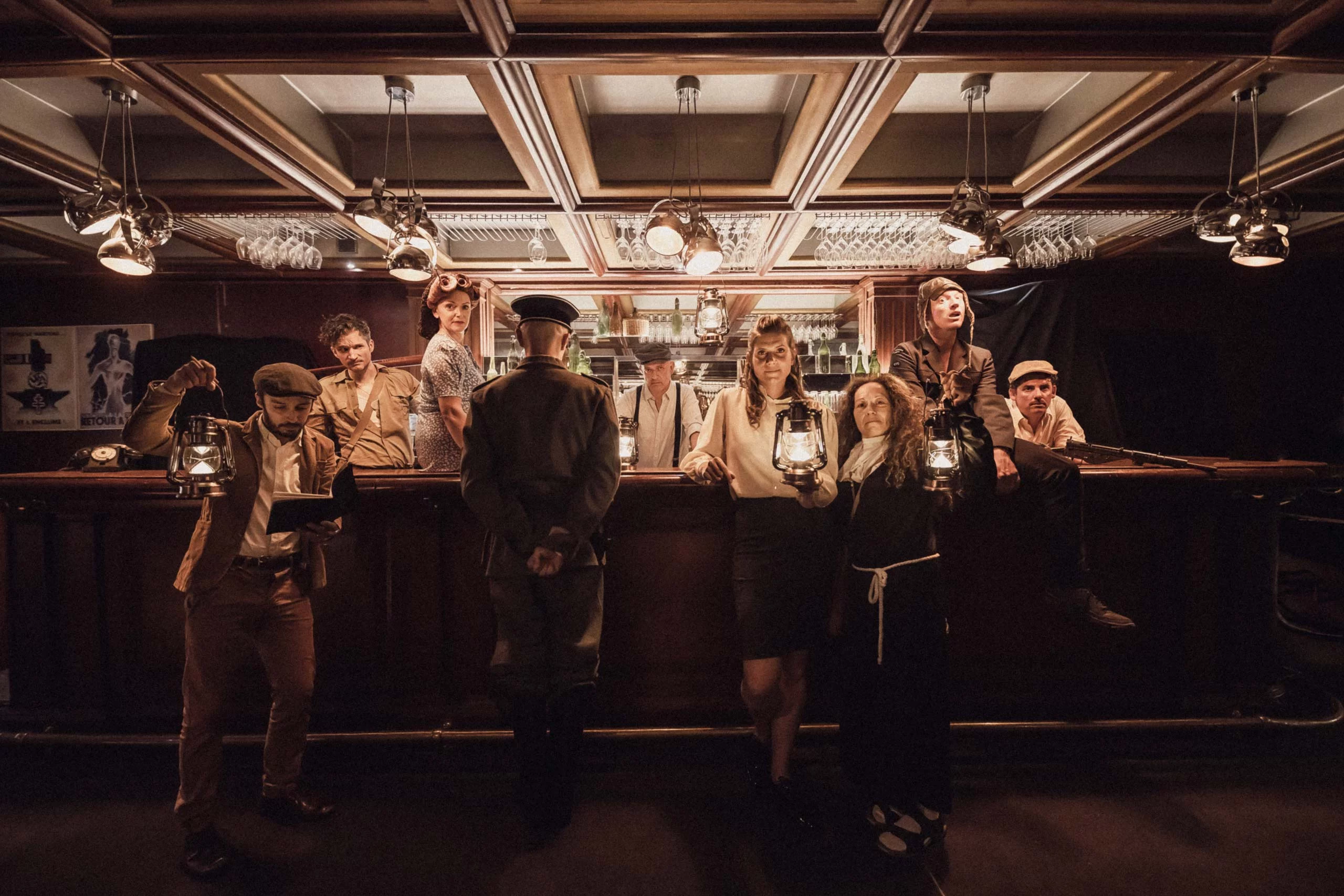 Group of nine people in vintage clothing posing inside a dimly lit bar, some holding lanterns and one man in military uniform.