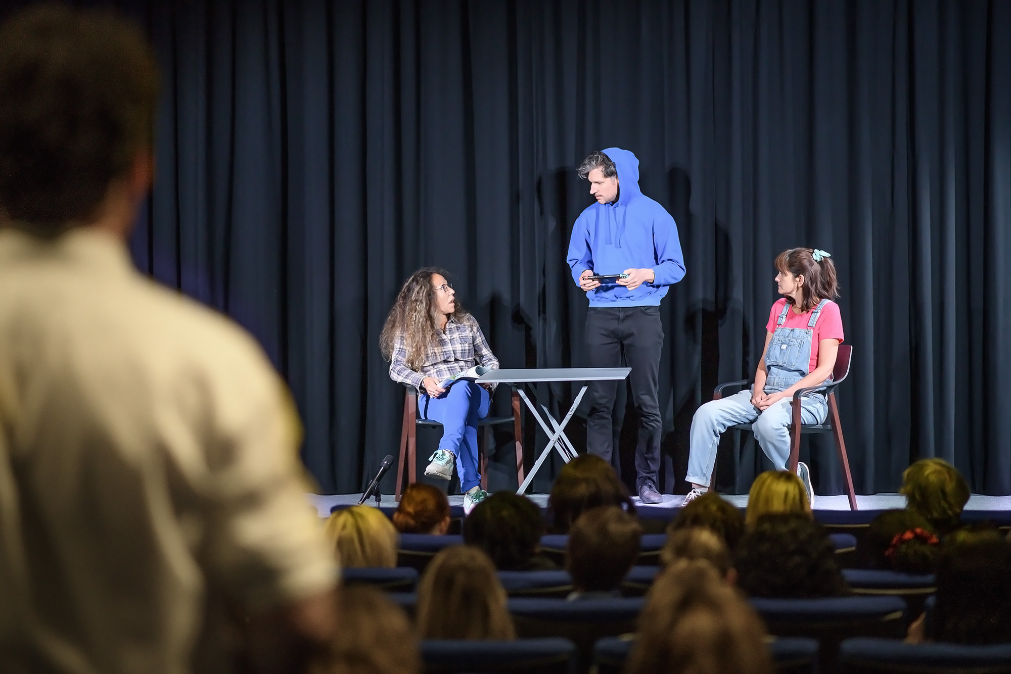 Trois acteurs en pleine scène sur scène avec un public assis devant eux dans un théâtre.