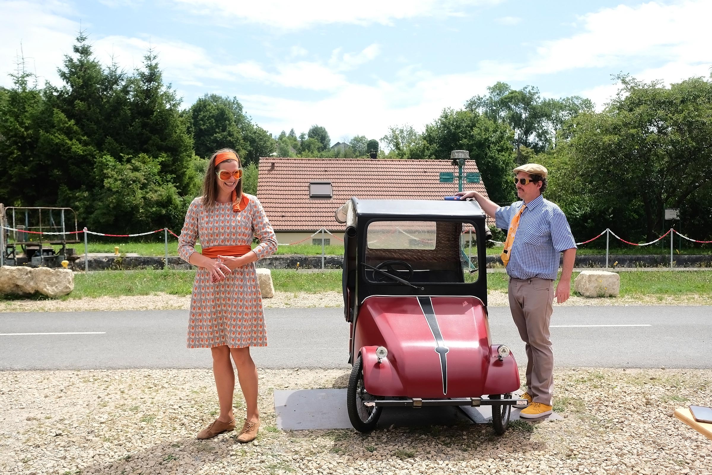 Un homme et une femme habillés vintage posent de chaque côté d’une petite voiture rouge avec une maison et des arbres en arrière-plan.