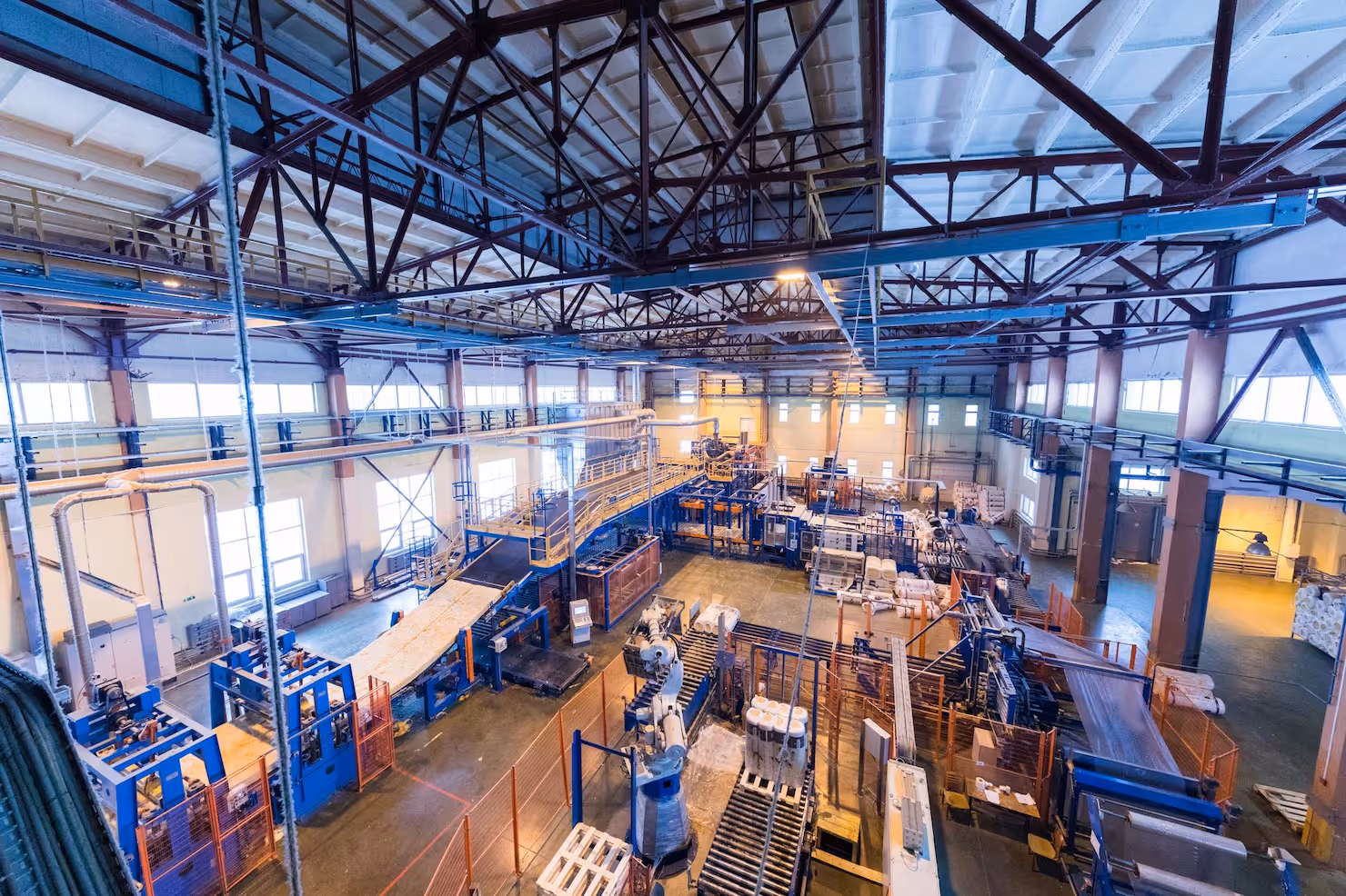 Wide view of an industrial factory interior with conveyor belts, machinery, and storage pallets under a high ceiling with metal trusses.