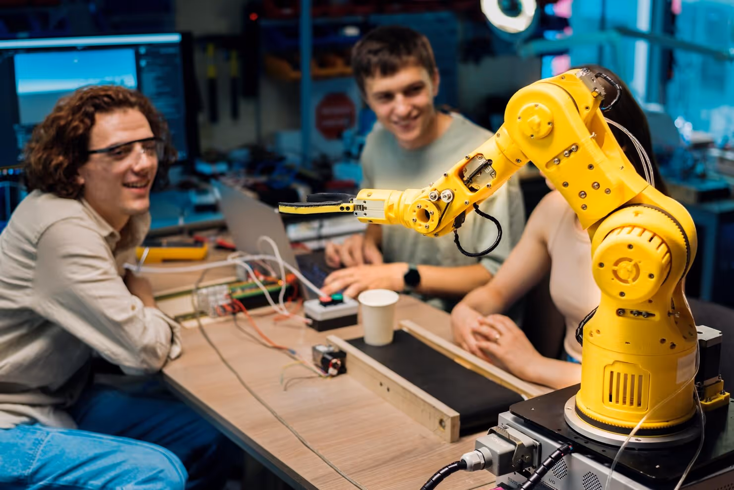 Three young people sit around a table with a yellow robotic arm and electronic equipment in a tech workspace.