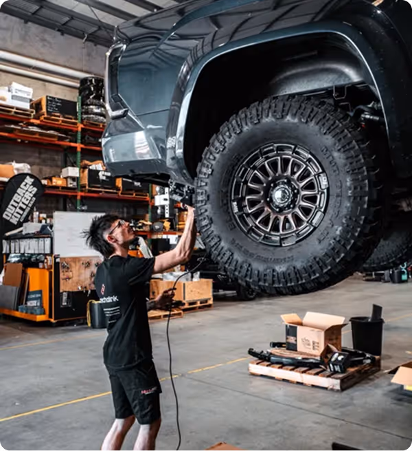 Mechanic working on a lifted off-road vehicle tire inside a garage with shelves and tools in the background.