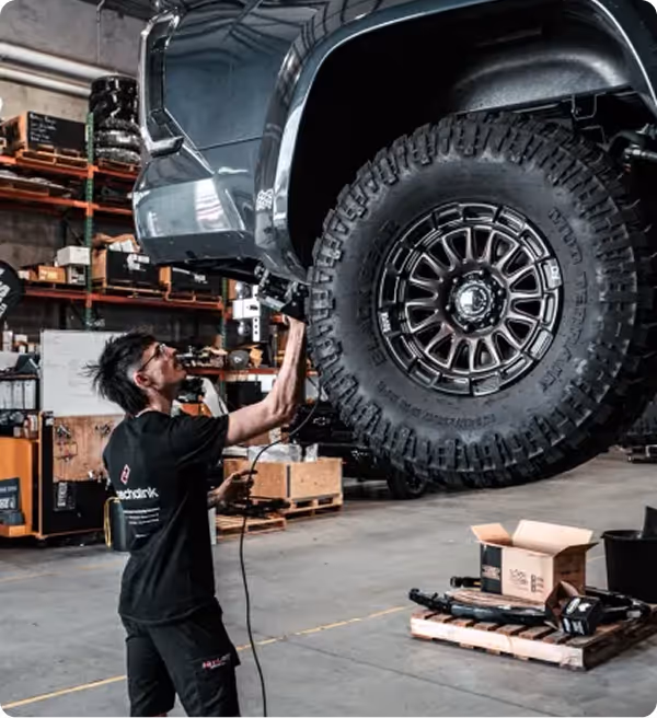 Mechanic working under the raised front end of a lifted off-road vehicle with large tires inside a garage.