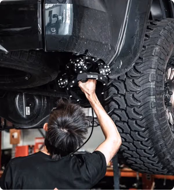 Person using a 3D scanner to inspect the underside of a raised vehicle with large tires in a workshop.