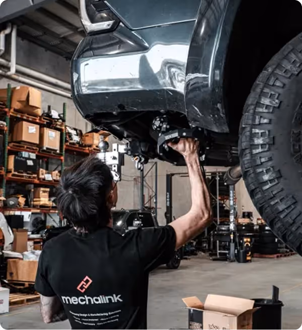 Mechanic working underneath a lifted vehicle in a workshop filled with shelves and automotive equipment.