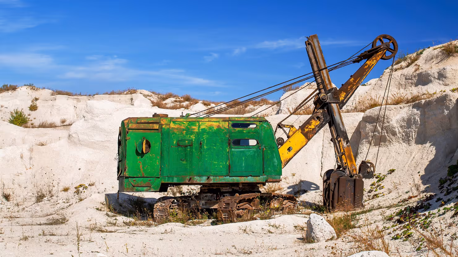 Rusty old green and yellow excavator with a pickaxe attachment in a white sandy quarry under a blue sky.