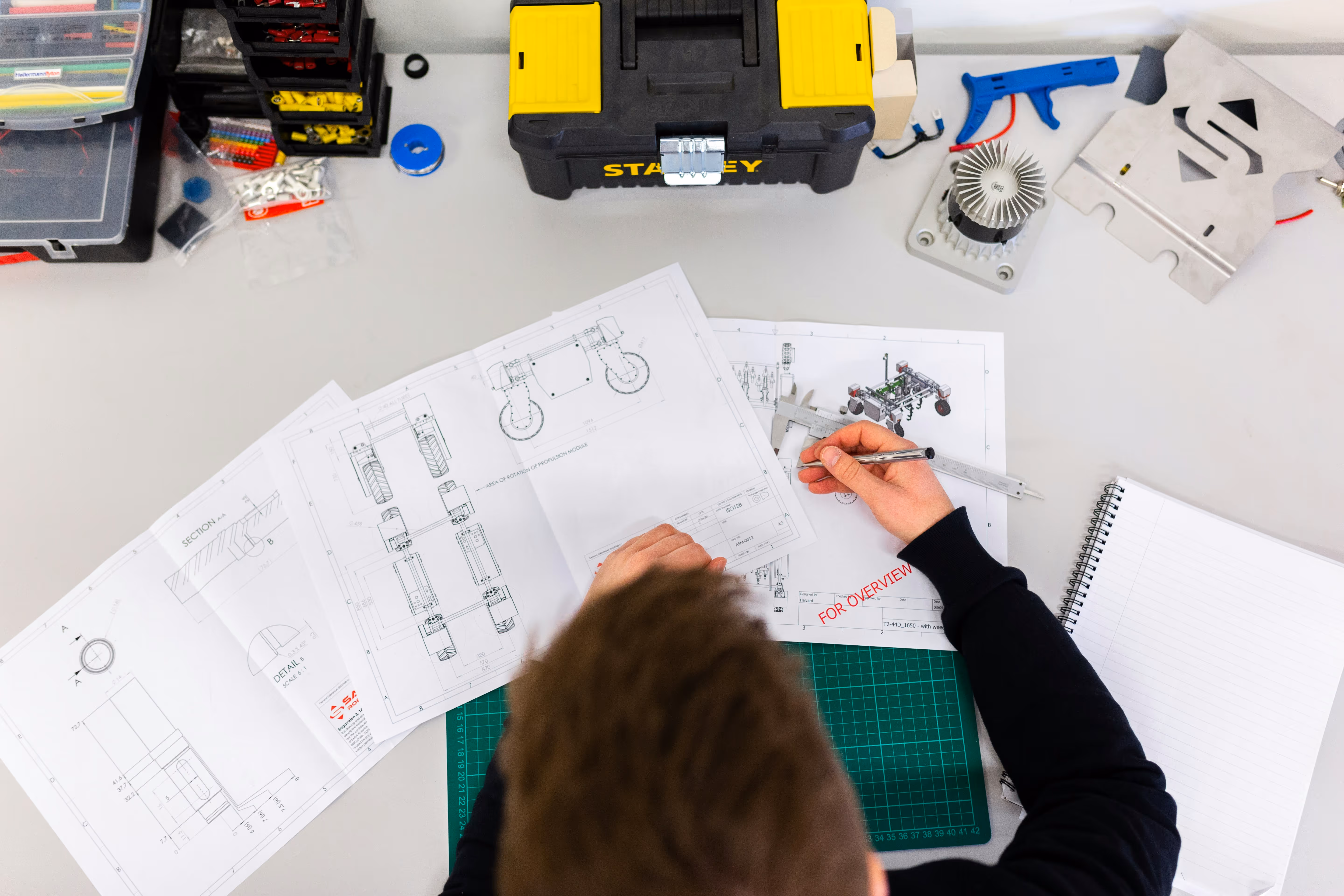Person working on technical blueprints with a caliper on a white desk surrounded by tools and a notebook.