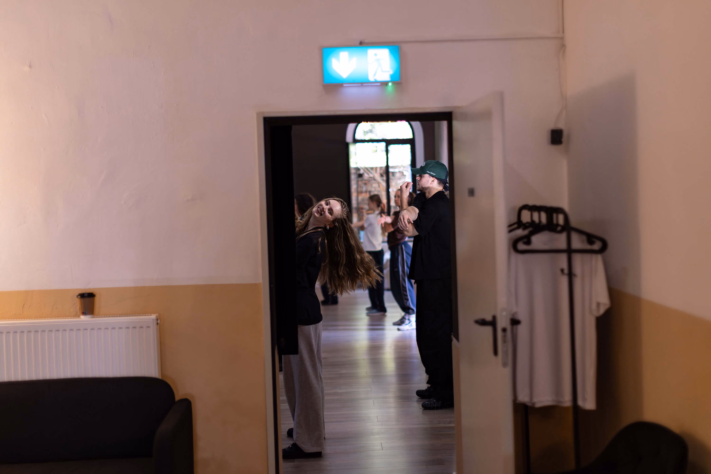 Frau lehnt sich spielerisch durch eine Tür, während mehrere Personen im Saal der Tanzschule The Base Erfurt im Hintergrund tanzen.