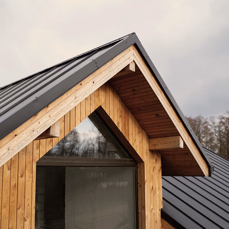 Modern wooden house roof detail with sloped metal roofing, exposed timber beams, and large triangular glass window.