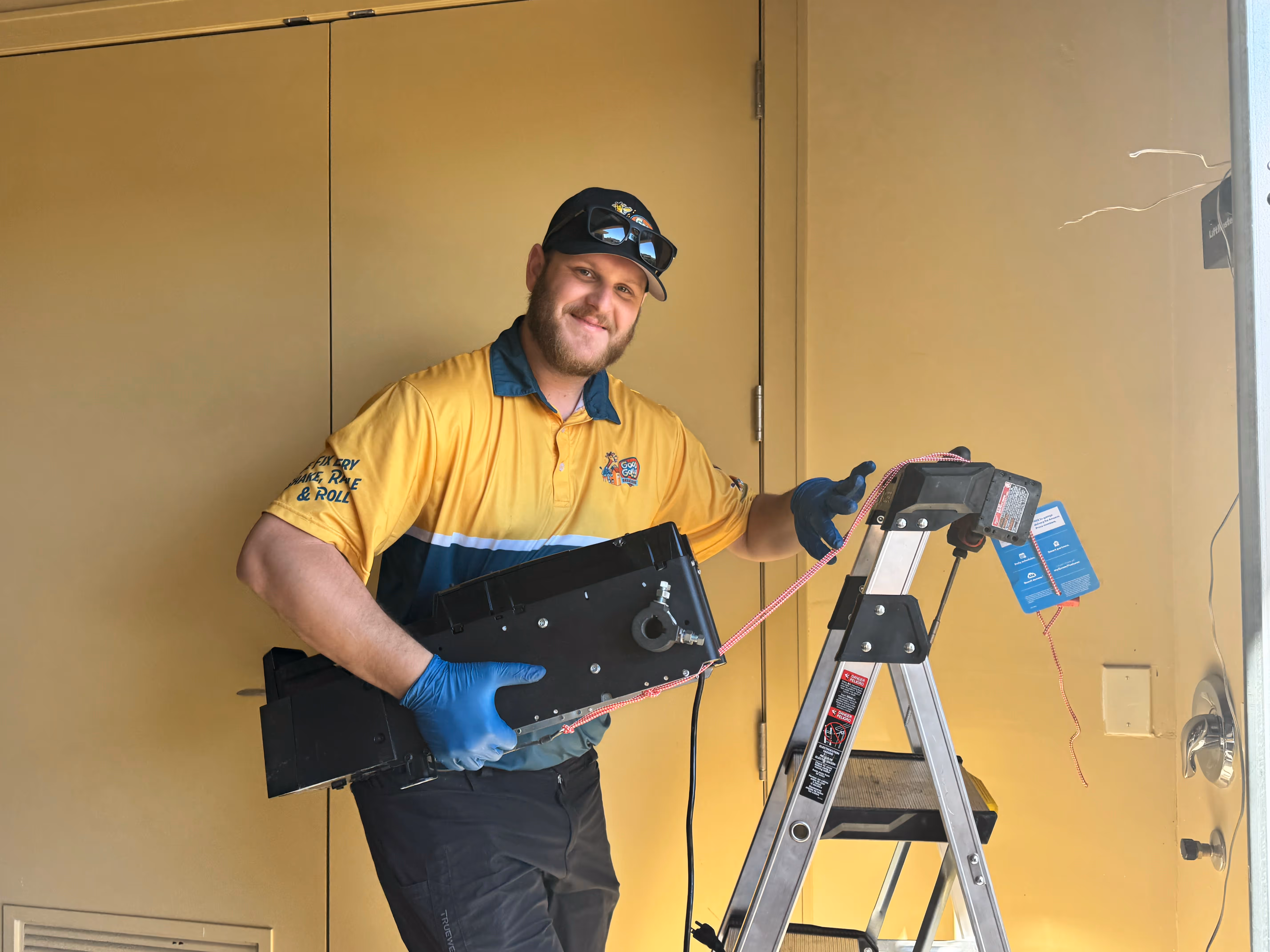 A man wearing a yellow and blue uniform and a cap, standing on a ladder, holds equipment while smiling.