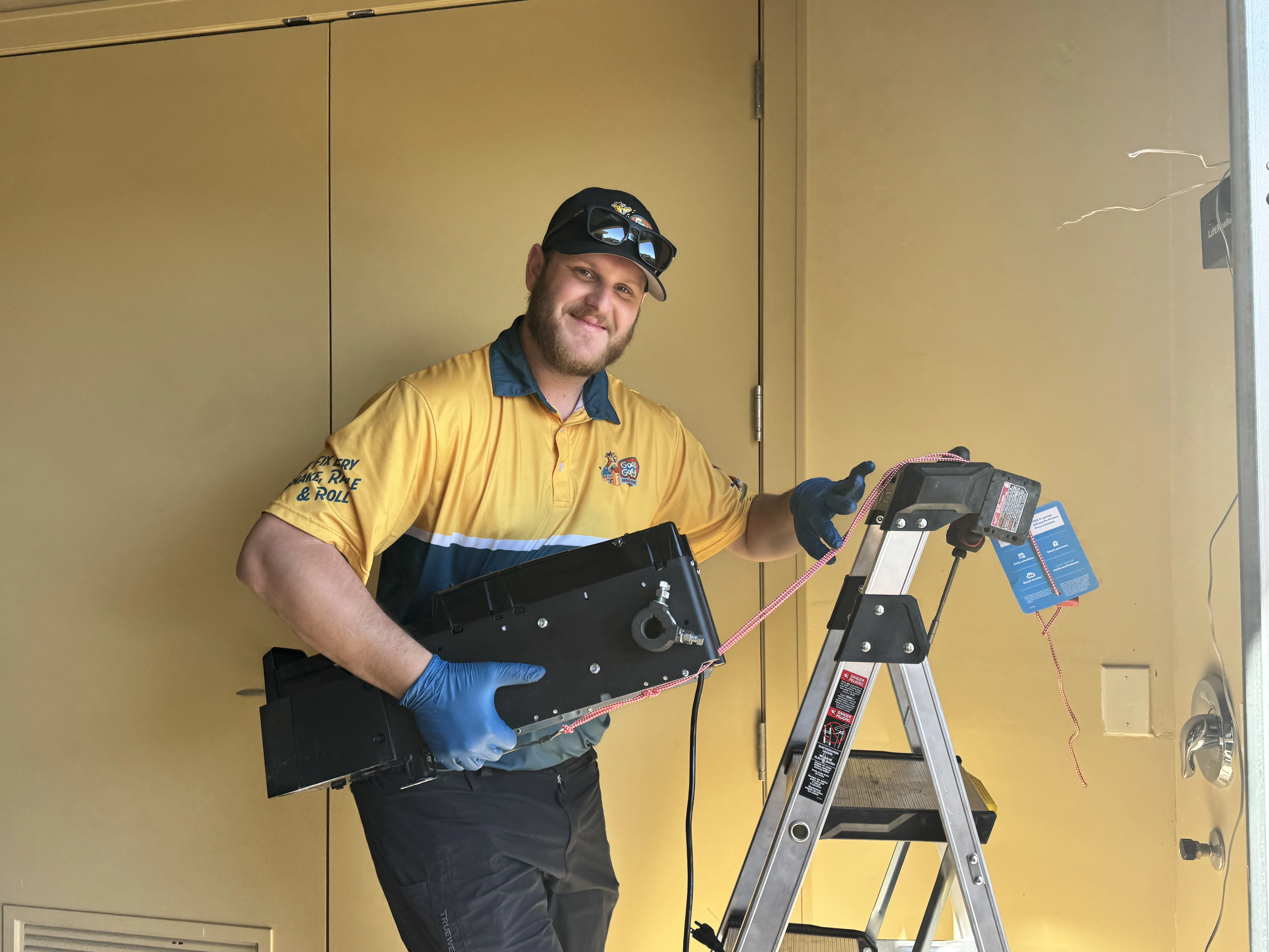 A man wearing a yellow and blue uniform and a cap, standing on a ladder, holds equipment while smiling.