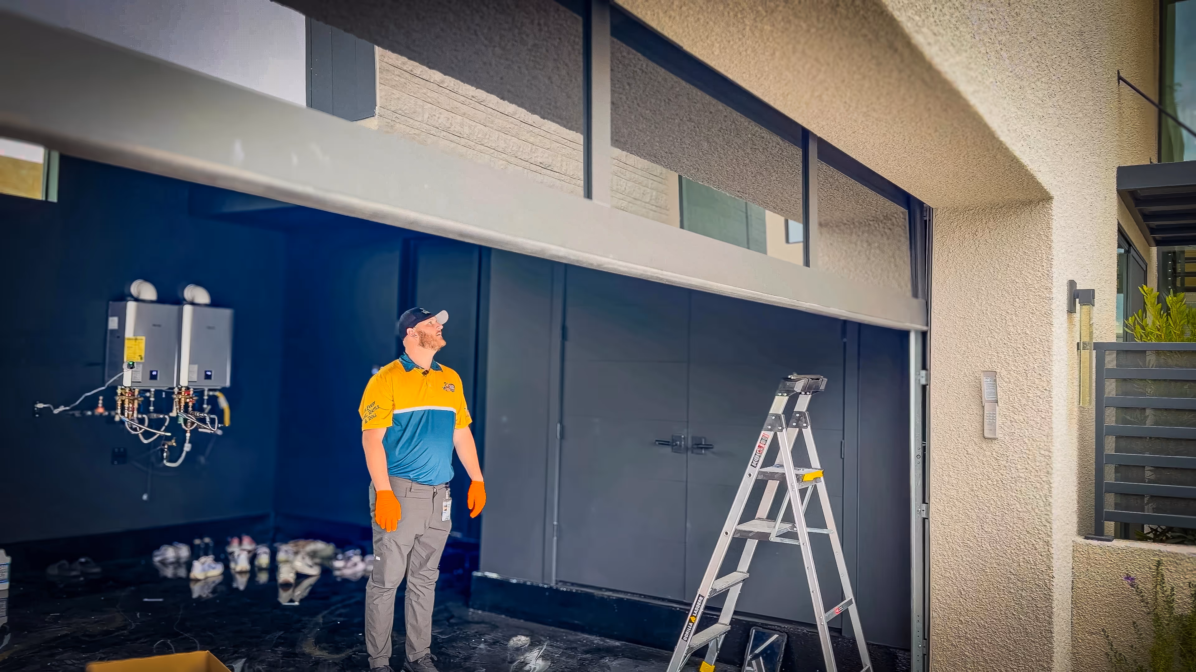 A man in a yellow and blue uniform inspects an open garage door. He wears orange gloves, with a ladder nearby. 