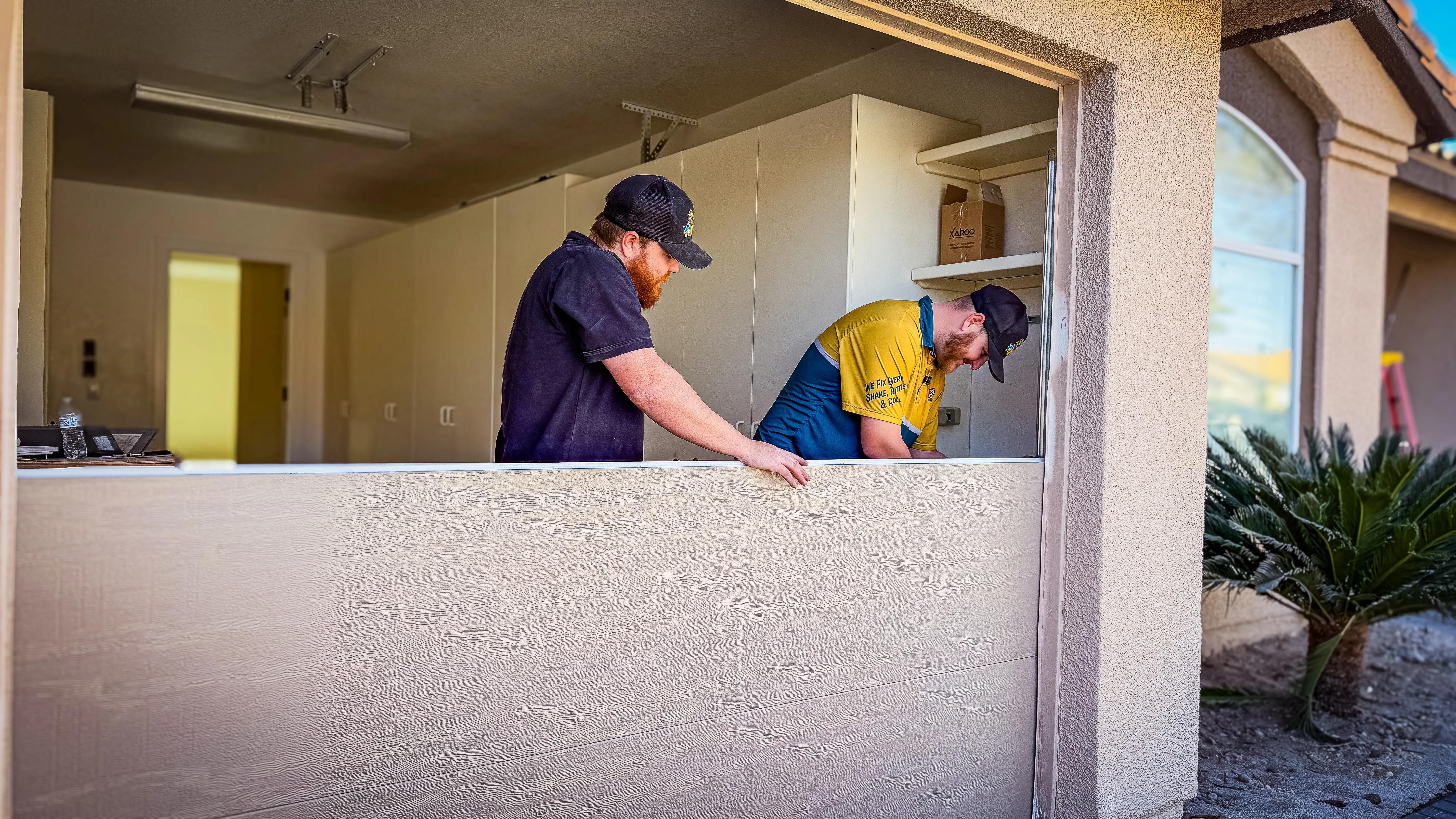 Two men in caps work together installing a large white board in a garage. 