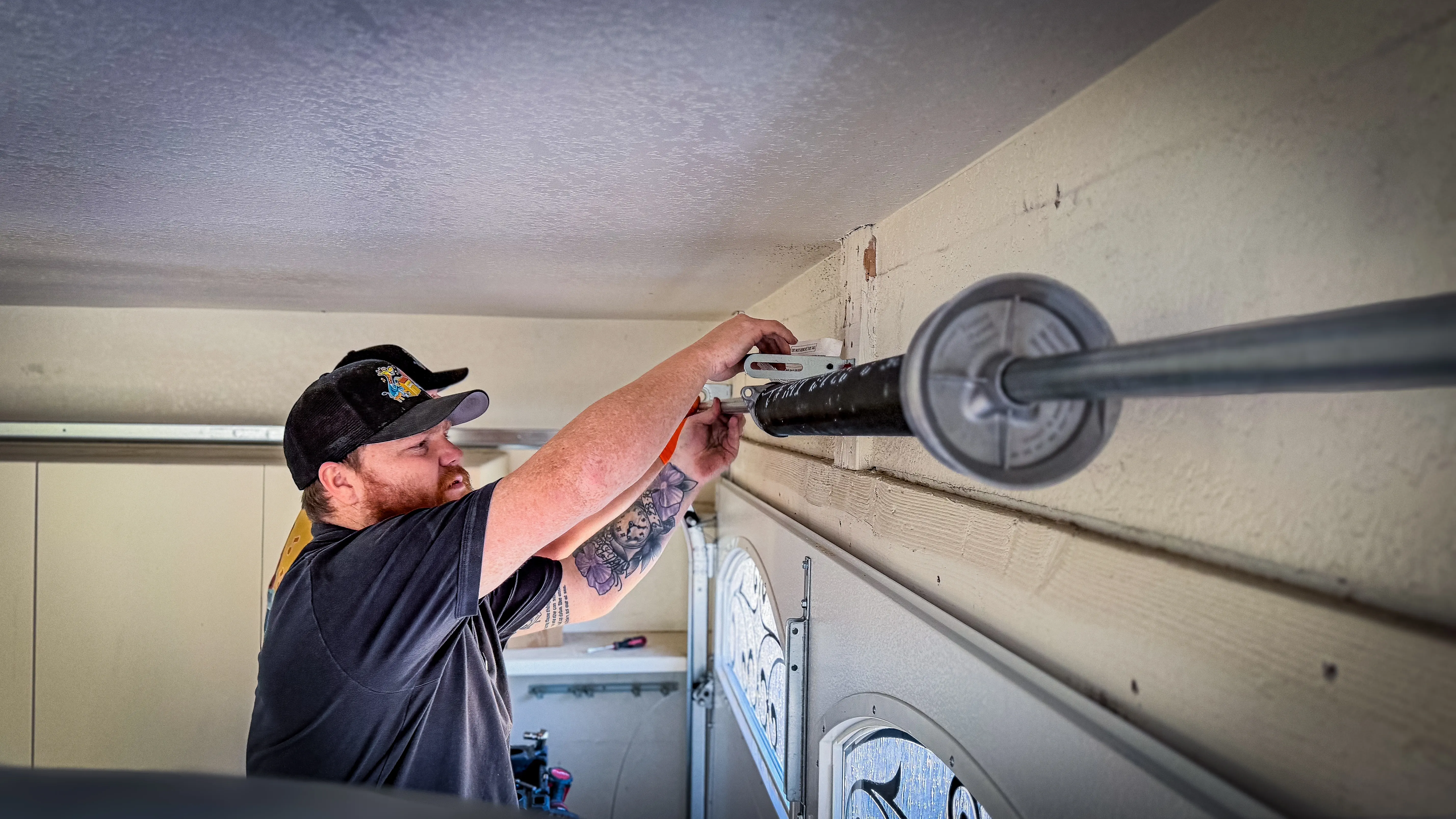 Man in a black cap and shirt repairs a garage door mechanism, focusing intently. 