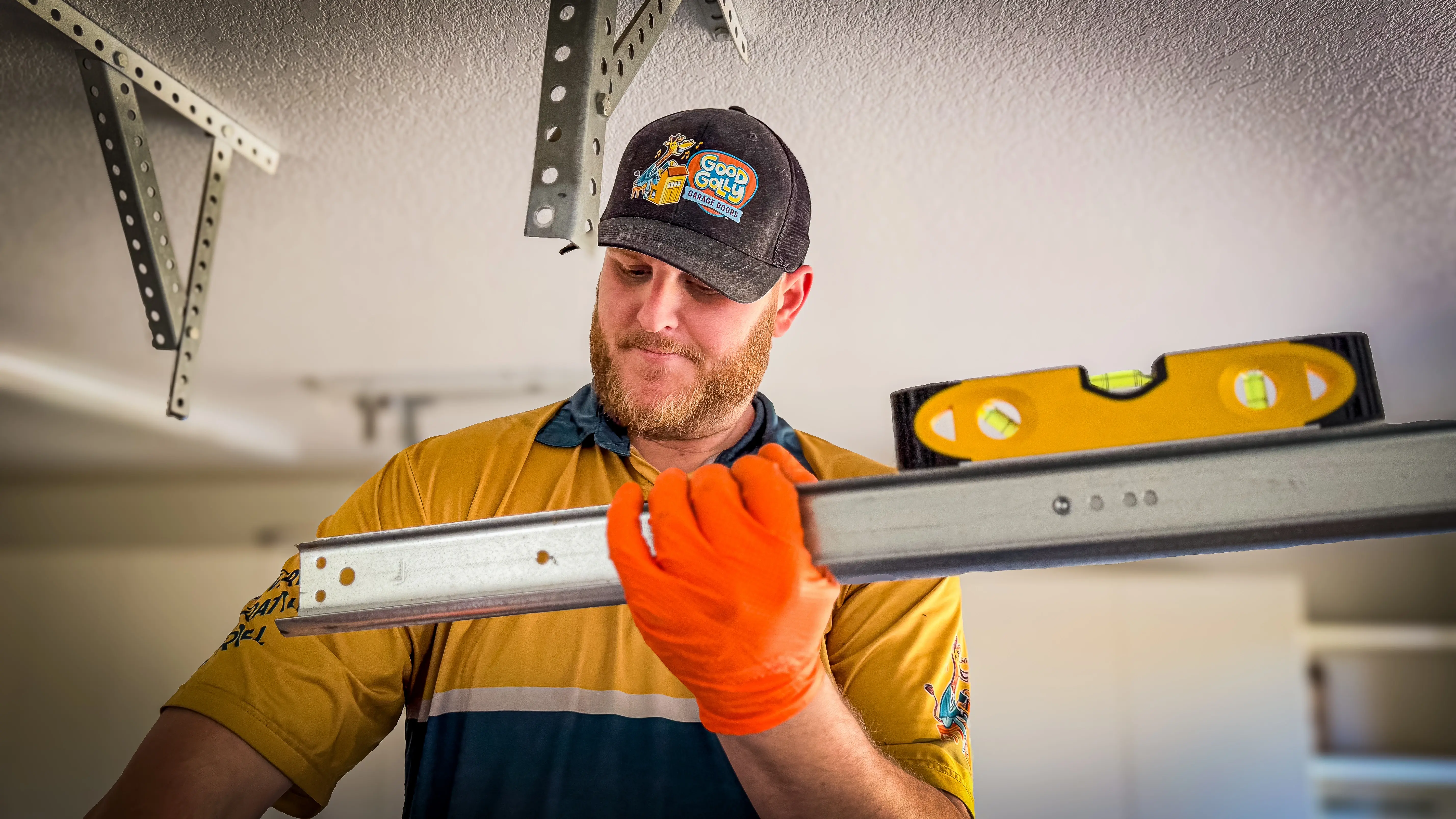 A man wearing an orange glove and a baseball cap holds a level and metal beam in a garage.