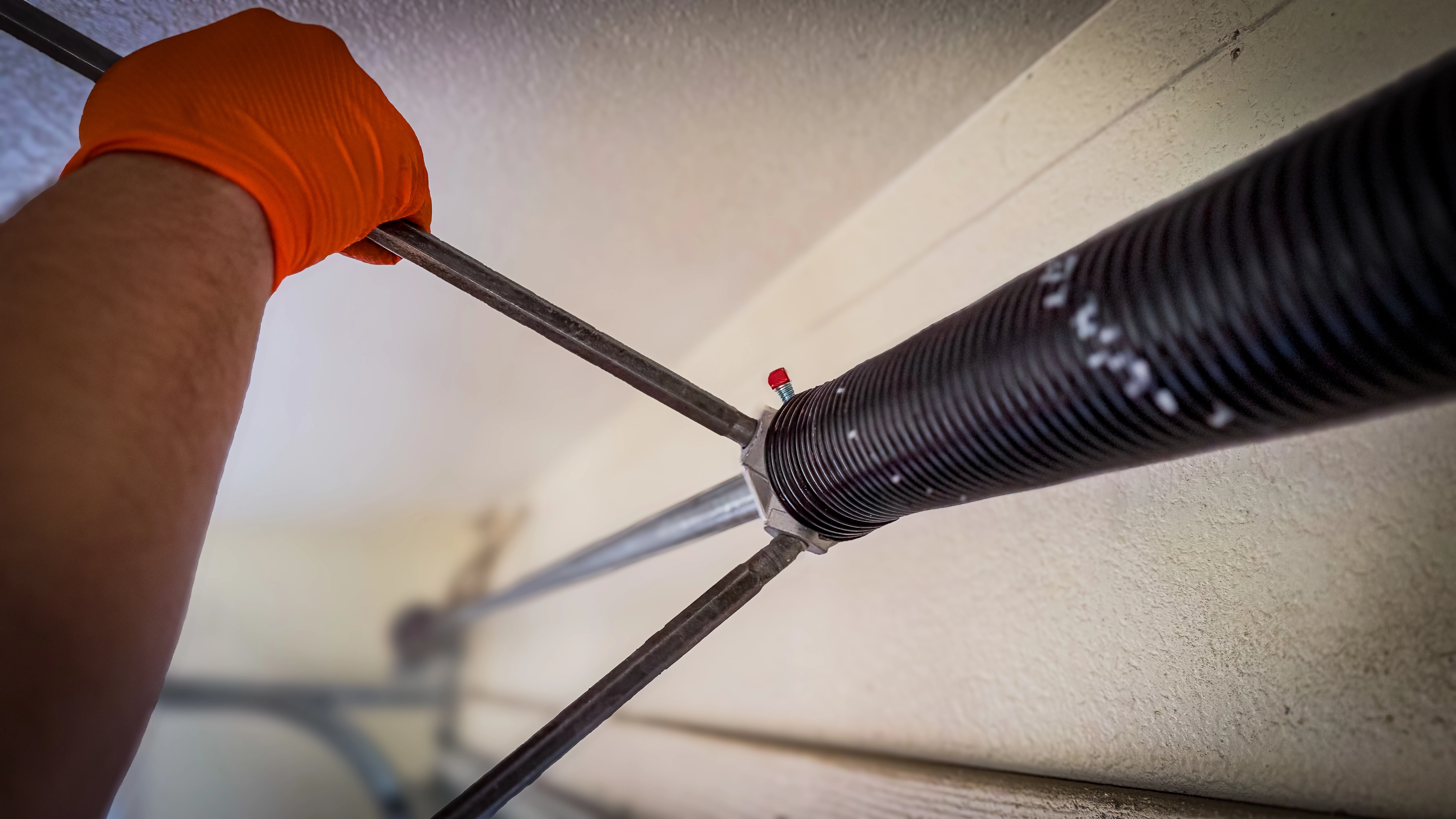 A person wearing an orange glove is adjusting a garage door torsion spring with a metal bar. 