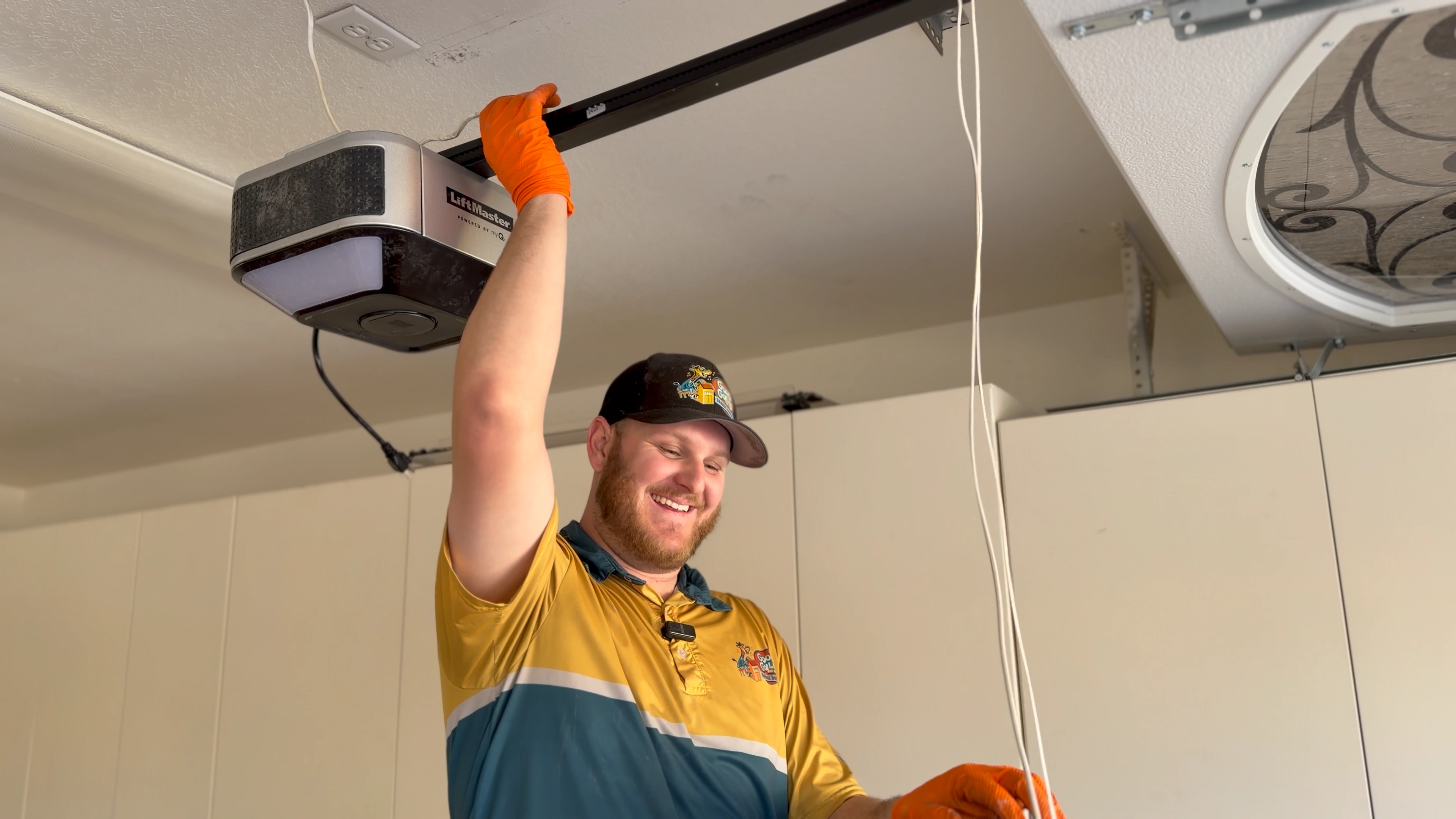A man wearing a cap and orange gloves is smiling while installing or repairing a garage door opener.