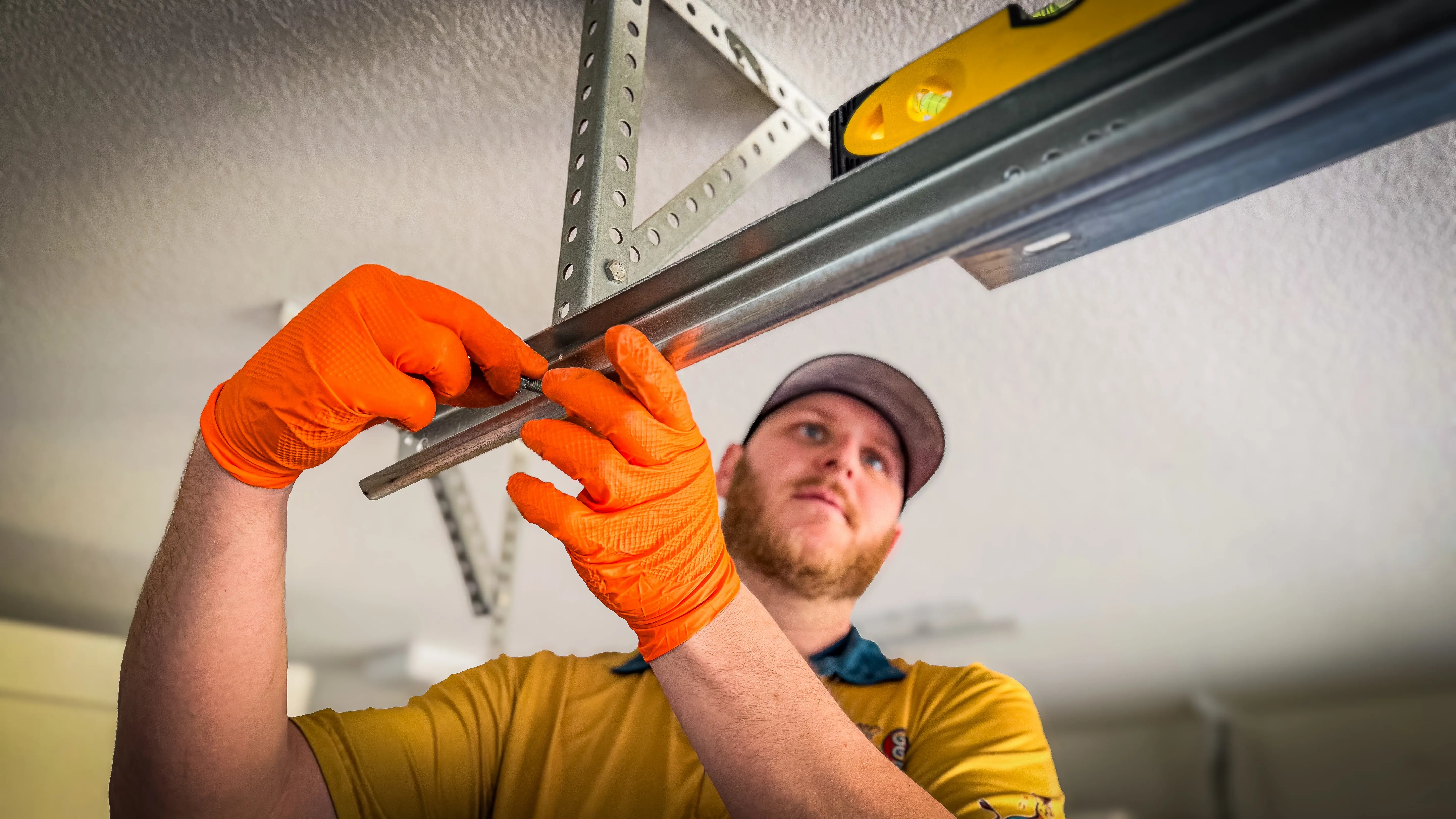 A person in a cap and orange gloves installs a metal beam on a ceiling, using a level for alignment.