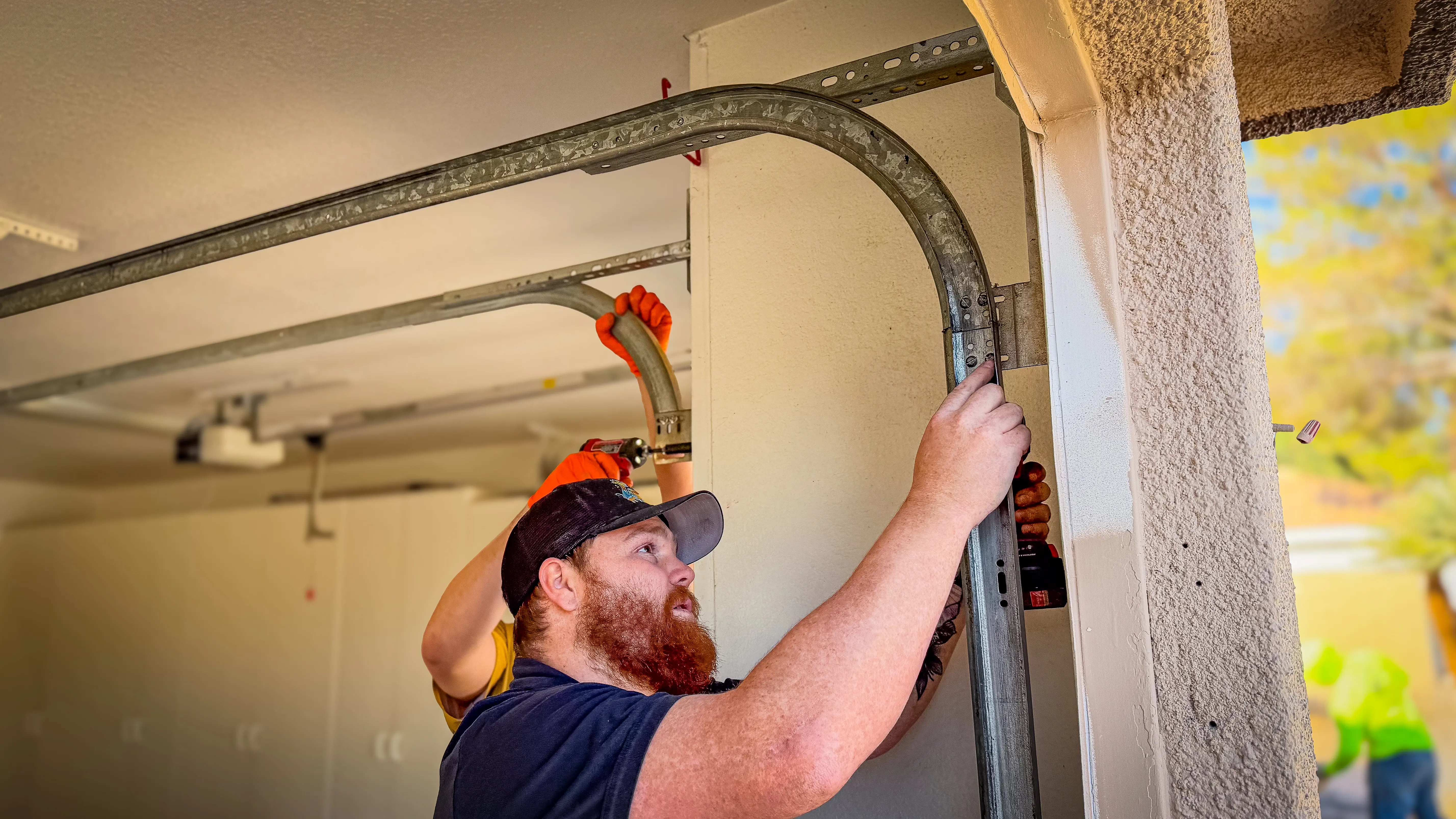 A man with a beard and cap installs a metal garage door track inside a garage. 