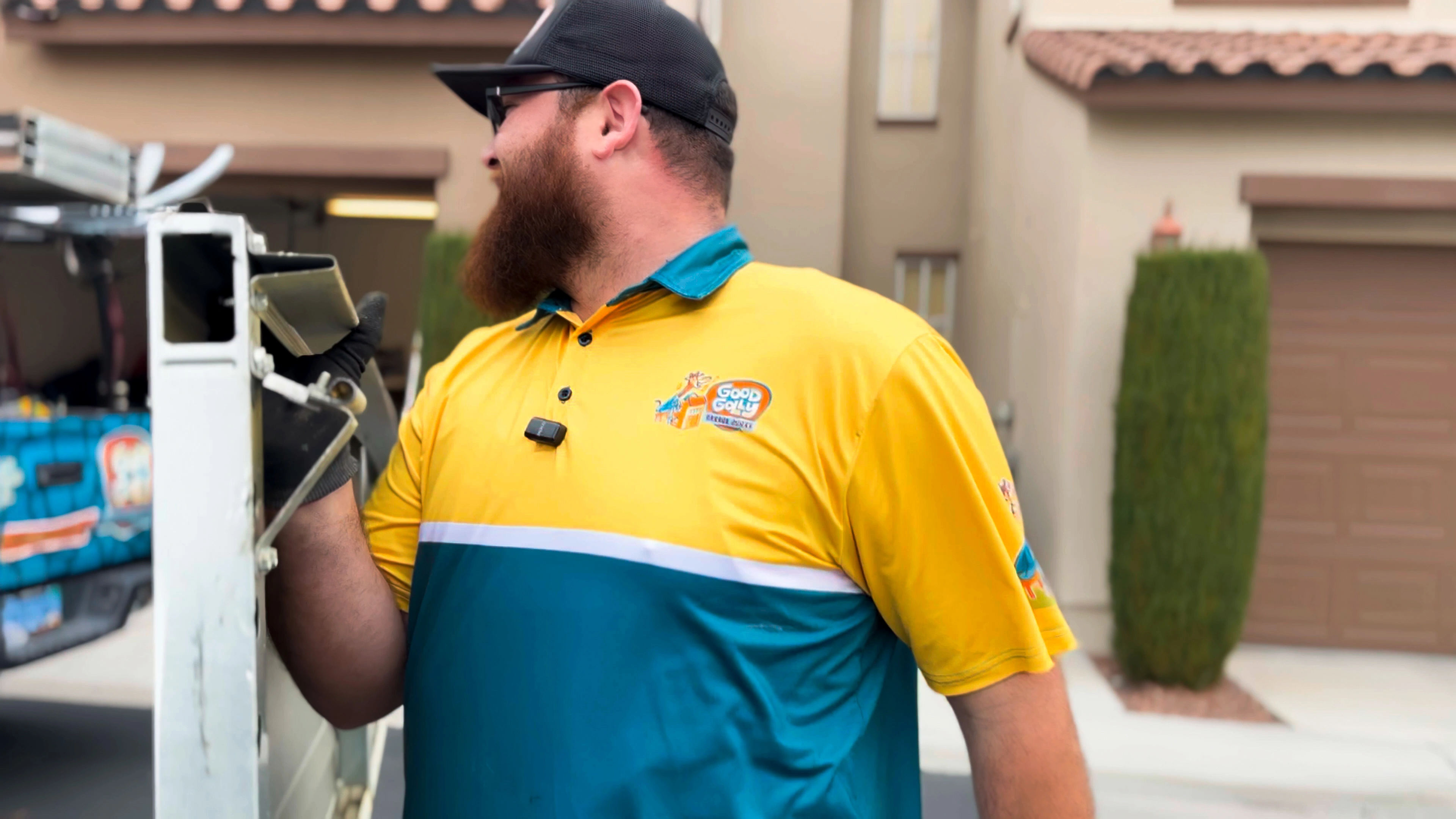 A man in a yellow and teal uniform with a logo is working outdoors. He is near a vehicle in a residential area