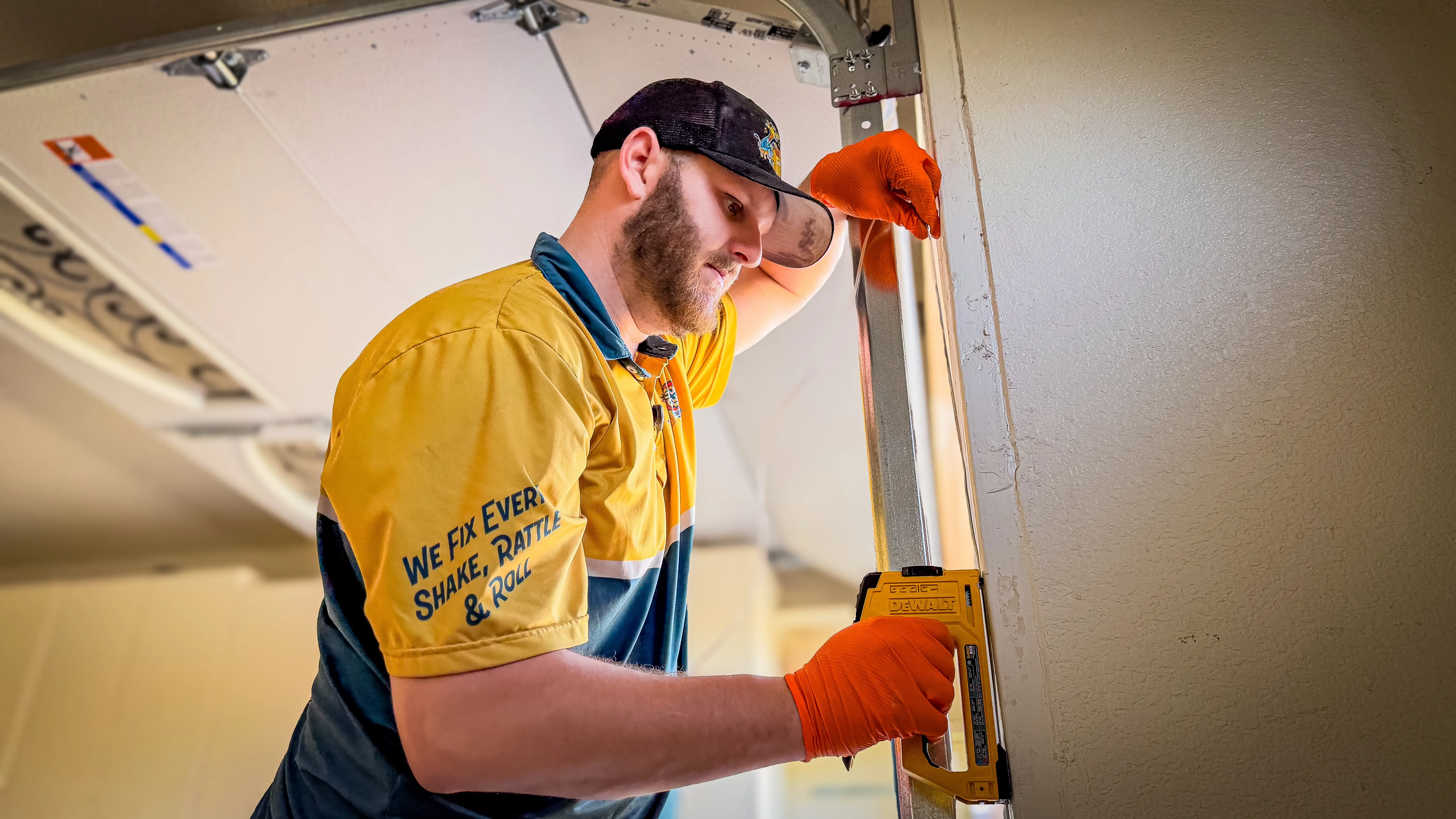 A man in a yellow and blue uniform, wearing orange gloves and a black cap, uses a power tool on a garage door.