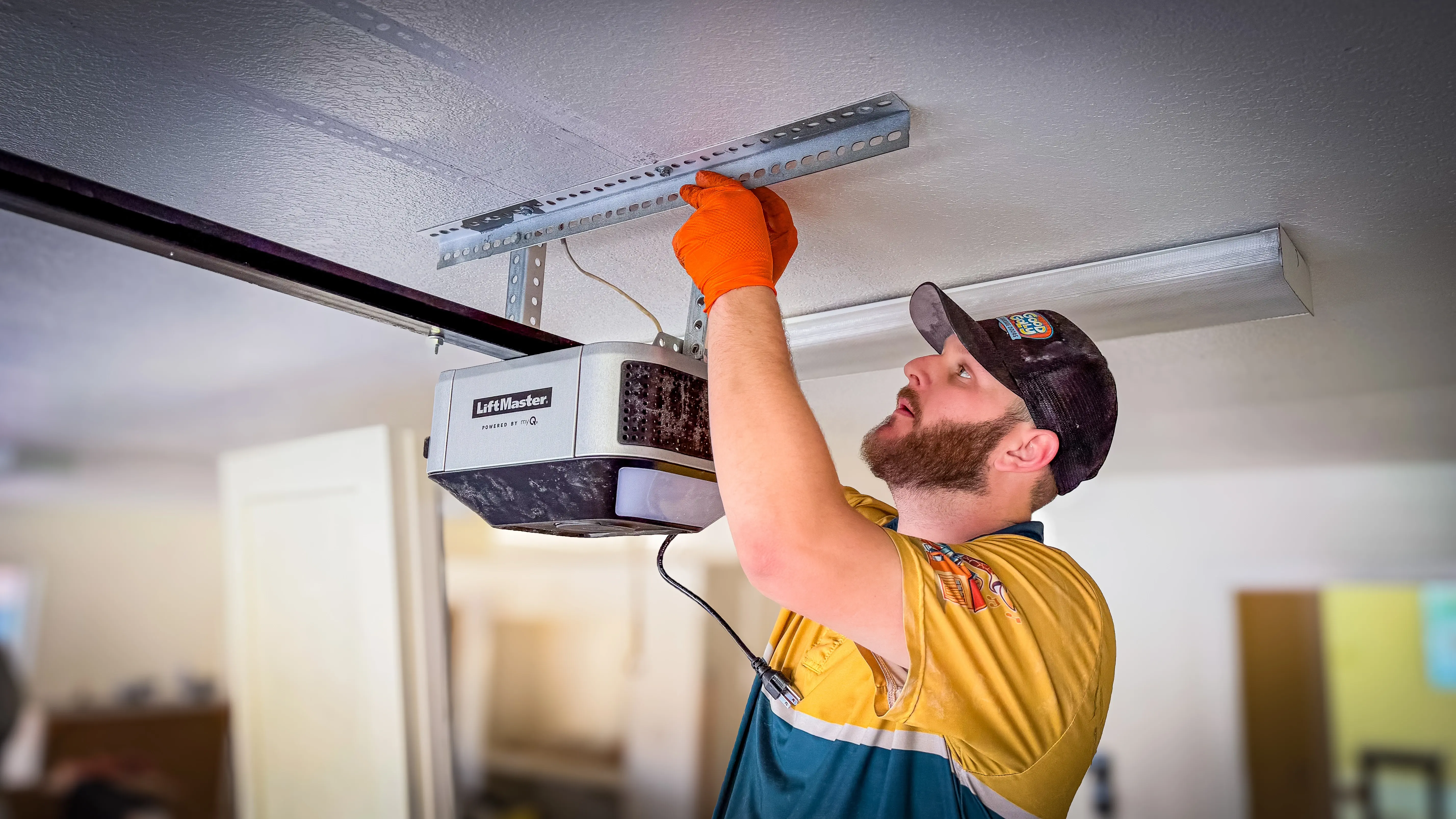 A man wearing an orange glove and work uniform installs a garage door opener on the ceiling, appearing focused and attentive. 