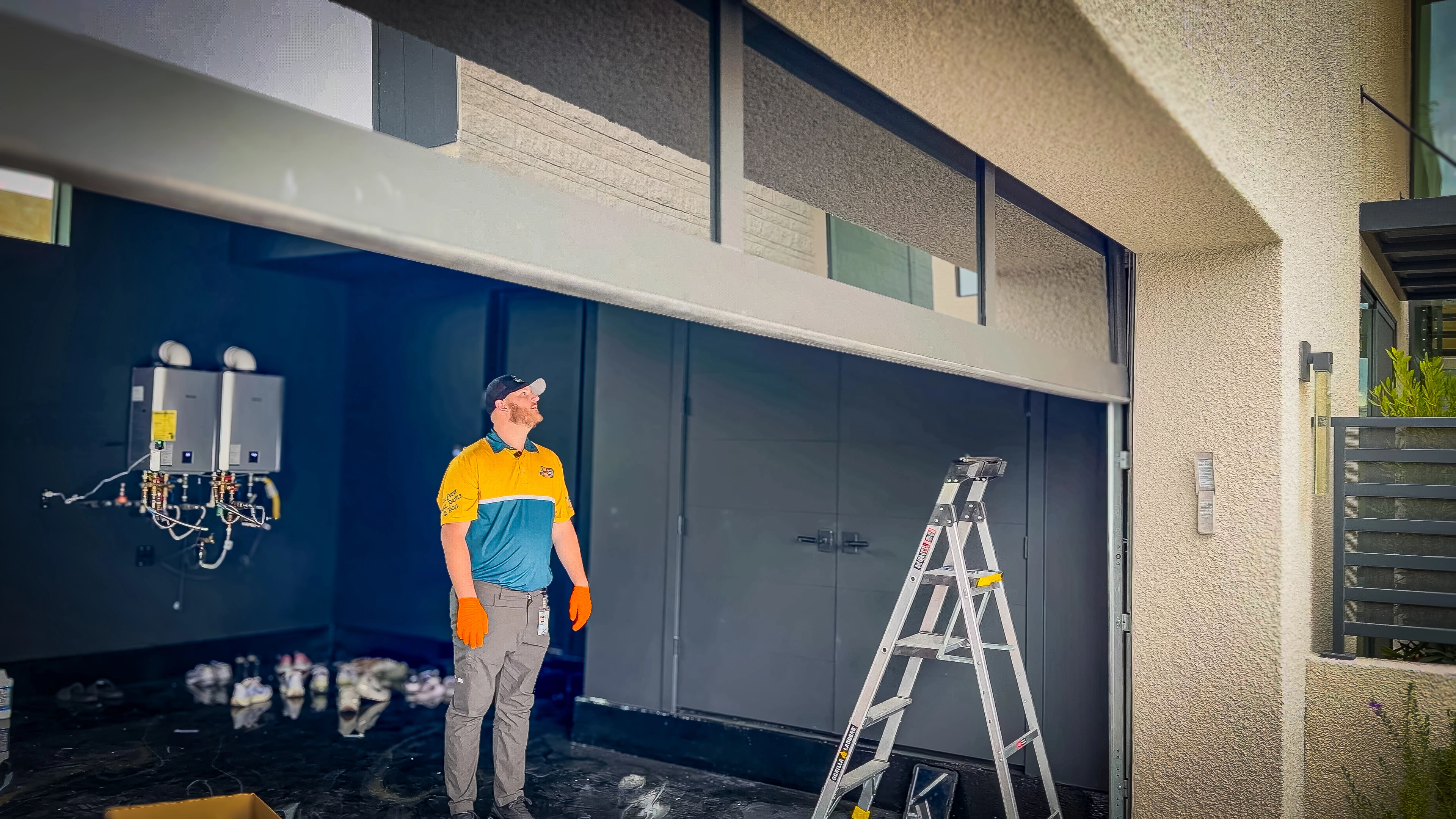 A man in a yellow and blue uniform inspects an open garage door. He wears orange gloves, with a ladder nearby.