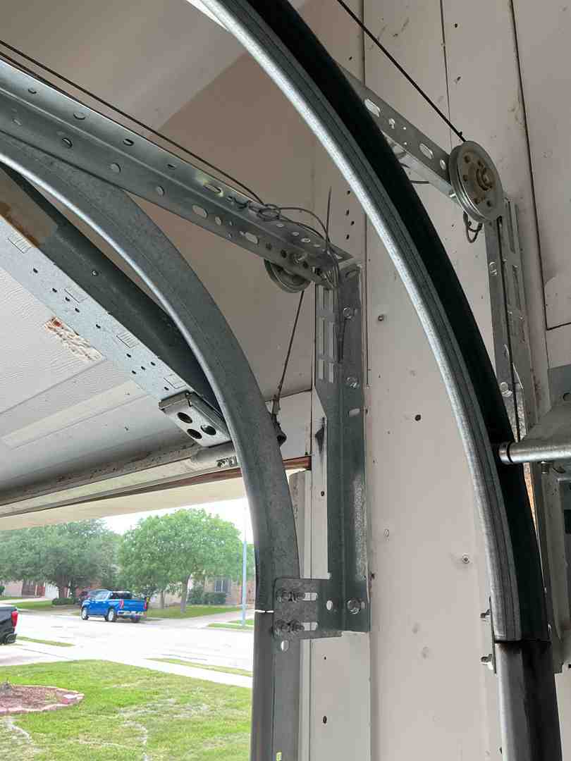 Infographic showing the anatomy of a garage door system with labeled components including vertical tracks on each side, horizontal rollers inside tracks, torsion springs mounted above the door, lift cables running from bottom brackets to drums, mounting brackets securing tracks to the wall, and the door opener mechanism - highlighting how each component must work in proper alignment to prevent the door from coming off track - Why Is My Garage Door Off Track In Georgetown TX