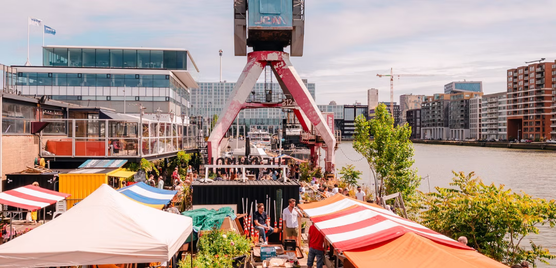 Levendige markt met kleurrijke kramen langs de waterkant in een stedelijk gebied met moderne gebouwen en een grote industriële kraan.