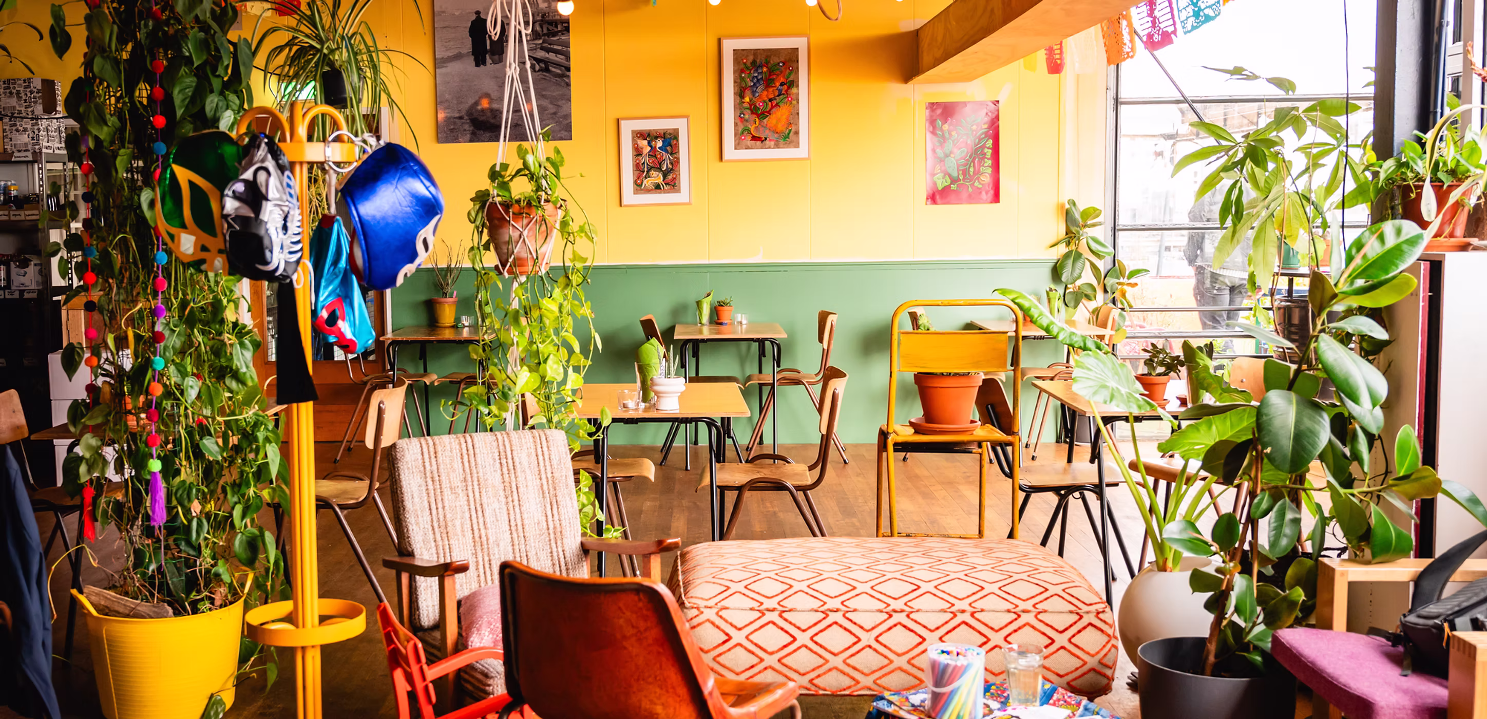 Gezellige café-inrichting met gele en groene muren, planten, verschillende stoelen, tafels en felgekleurde maskers aan een kapstok.