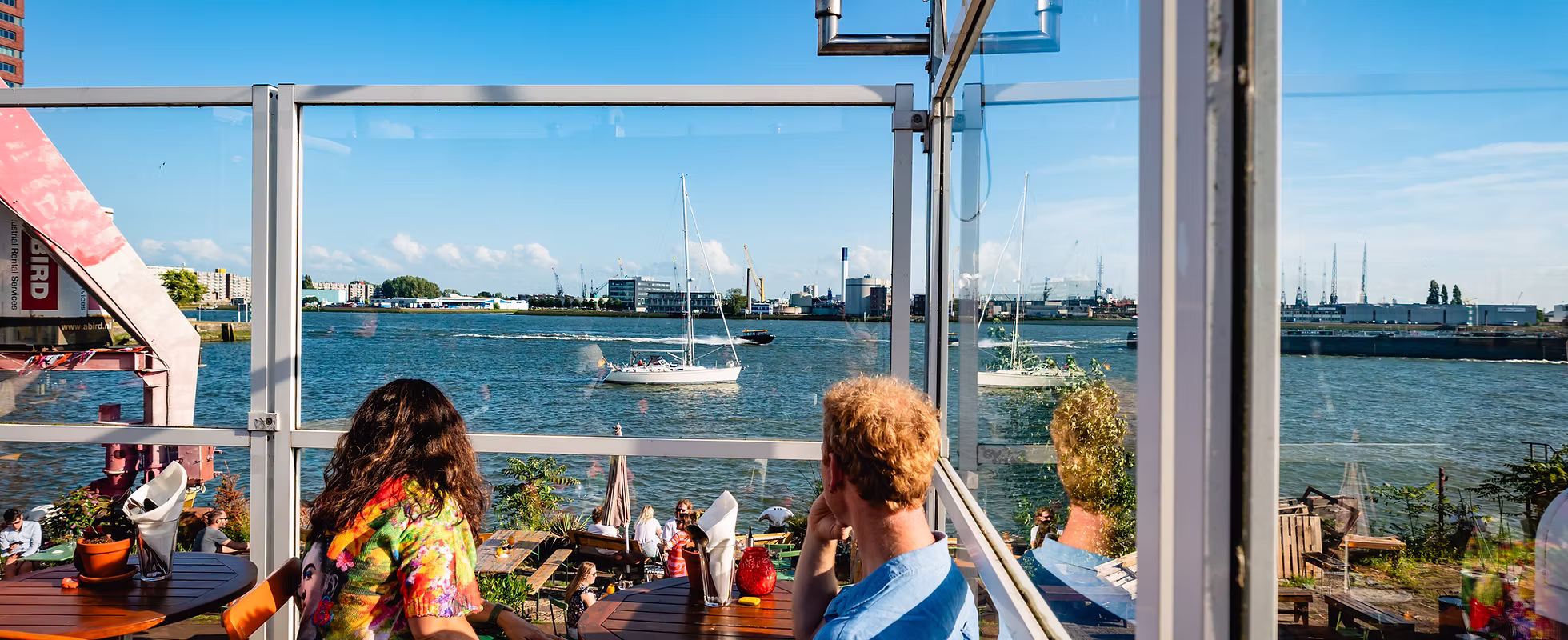 Twee mensen zitten aan een tafel met uitzicht op water, boten en een industriegebied aan de overkant onder een blauwe lucht.
