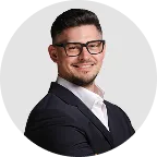 Lächelnder Mann mit kurzem dunklem Haar, Bart und Brille, trägt einen schwarzen Anzug und weißes Hemd, vor grauem Hintergrund.