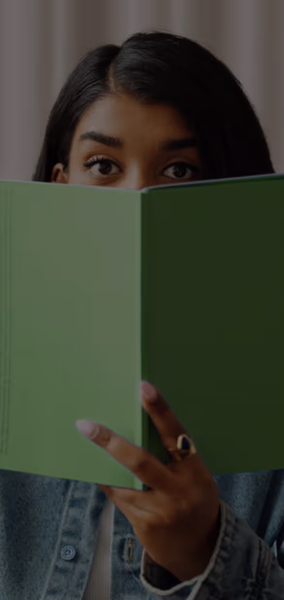 Person with dark hair partially hidden behind a green book, wearing a denim jacket and a ring.