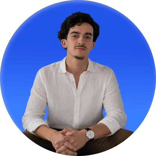 Man with dark curly hair wearing a white shirt and watch seated against a blue background.