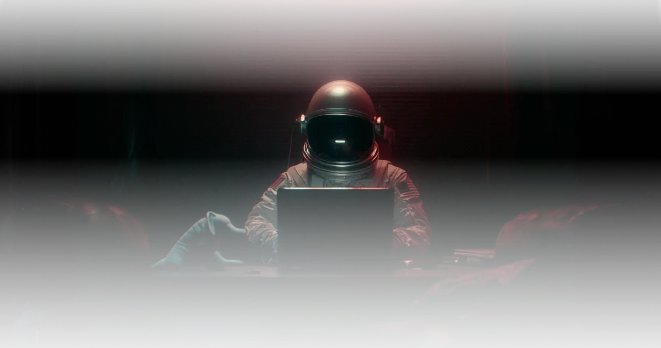 Astronaut sitting at a table with a laptop in a dimly lit room.