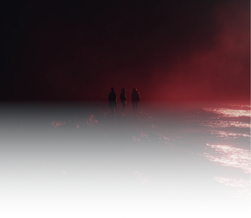 Three silhouetted figures standing on a rocky shore with dark red lighting and glowing ocean waves at night.
