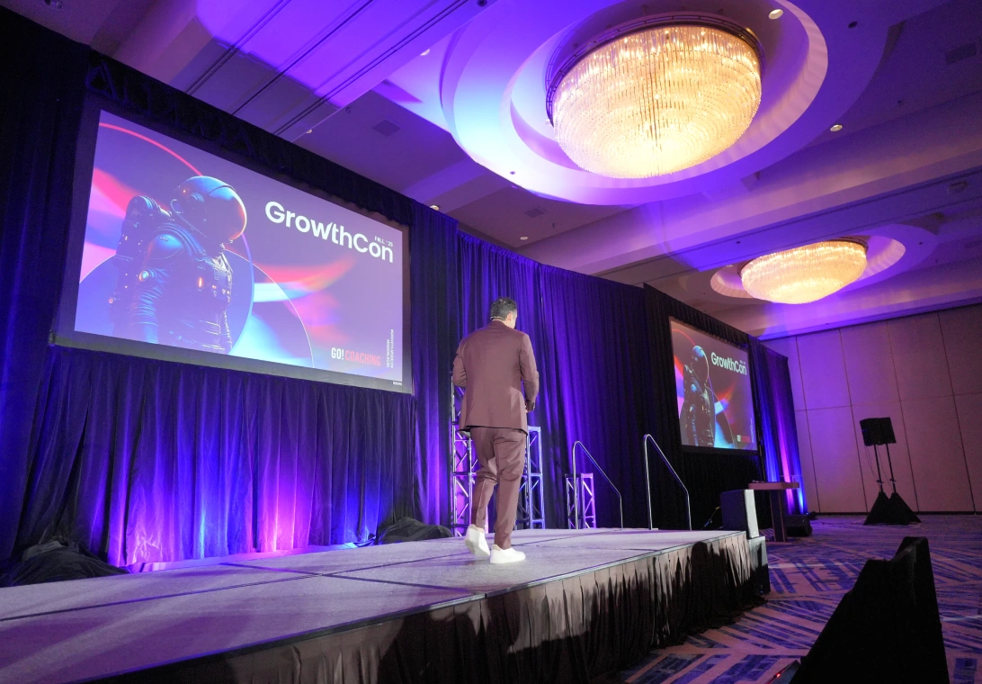 A man in a suit standing on a stage with two large screens displaying 'GrowthCon' and an astronaut image, under purple lighting in a conference room.