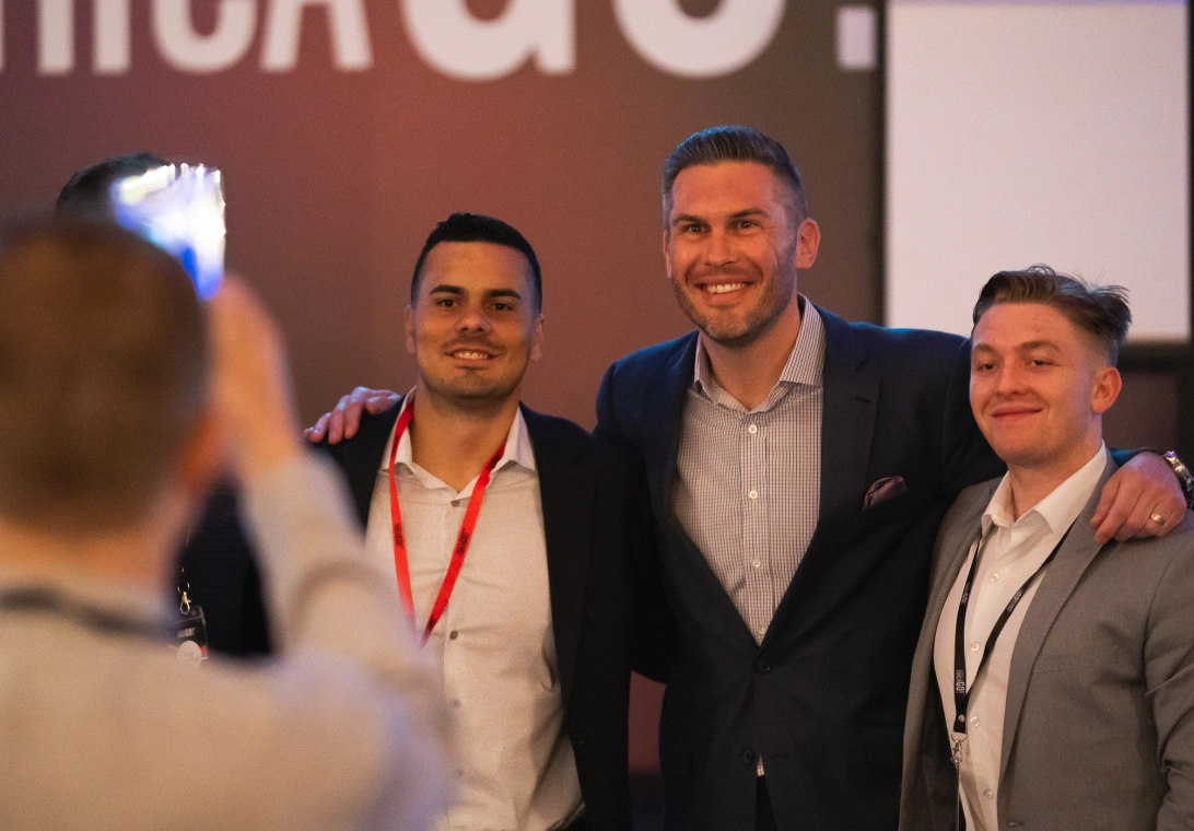 Three men in business attire smiling and posing for a photo taken by another person blurred in the foreground.